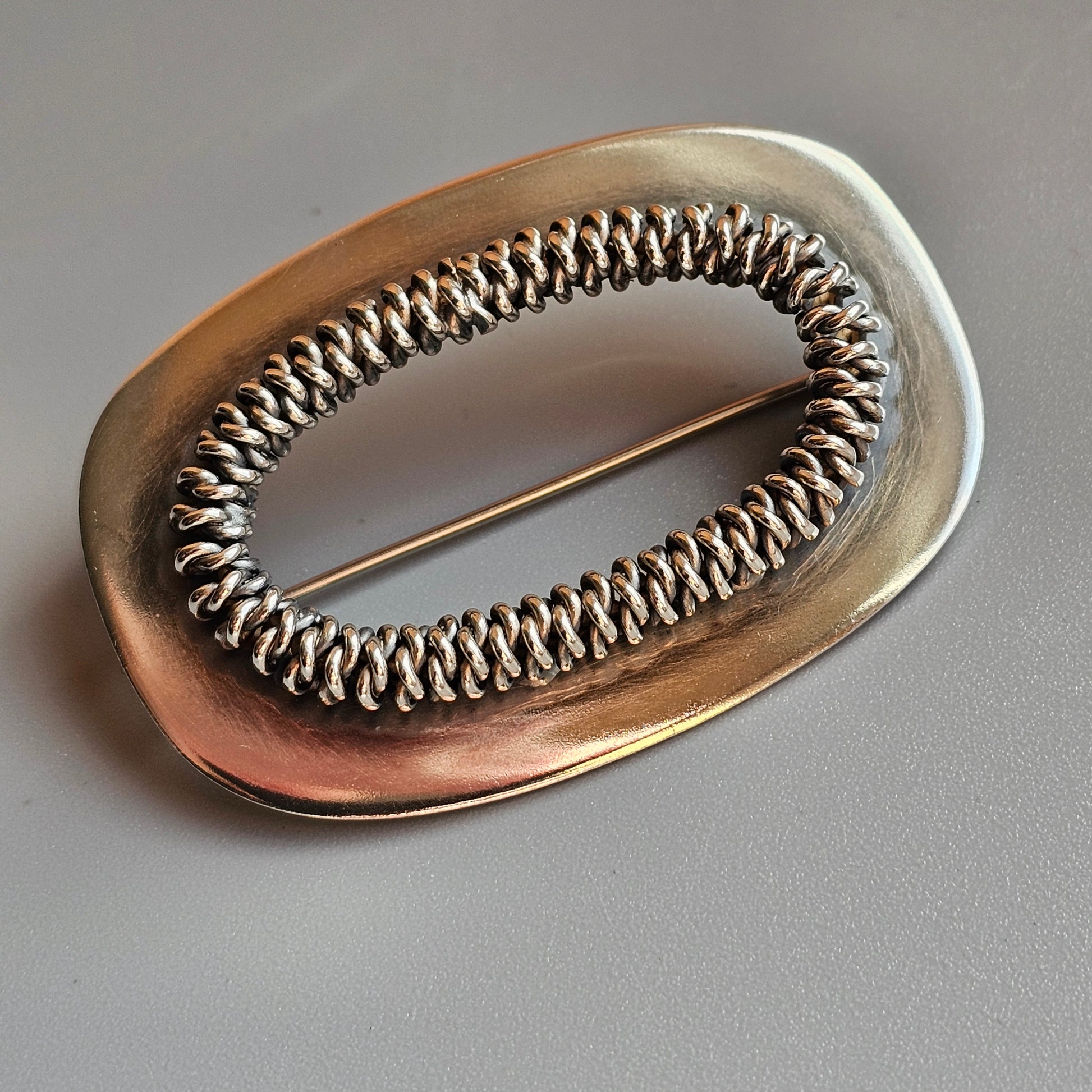 Oval-shaped metallic brooch with an intricate spiral wire design in the center.