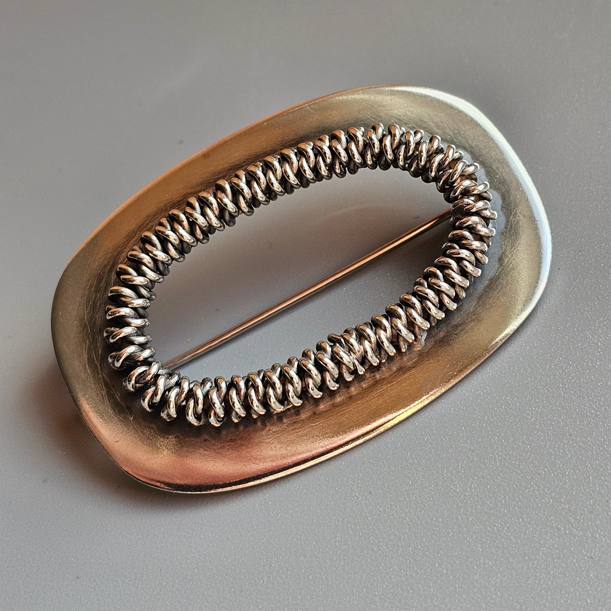 Oval-shaped metallic brooch with an intricate coiled wire design in the center.