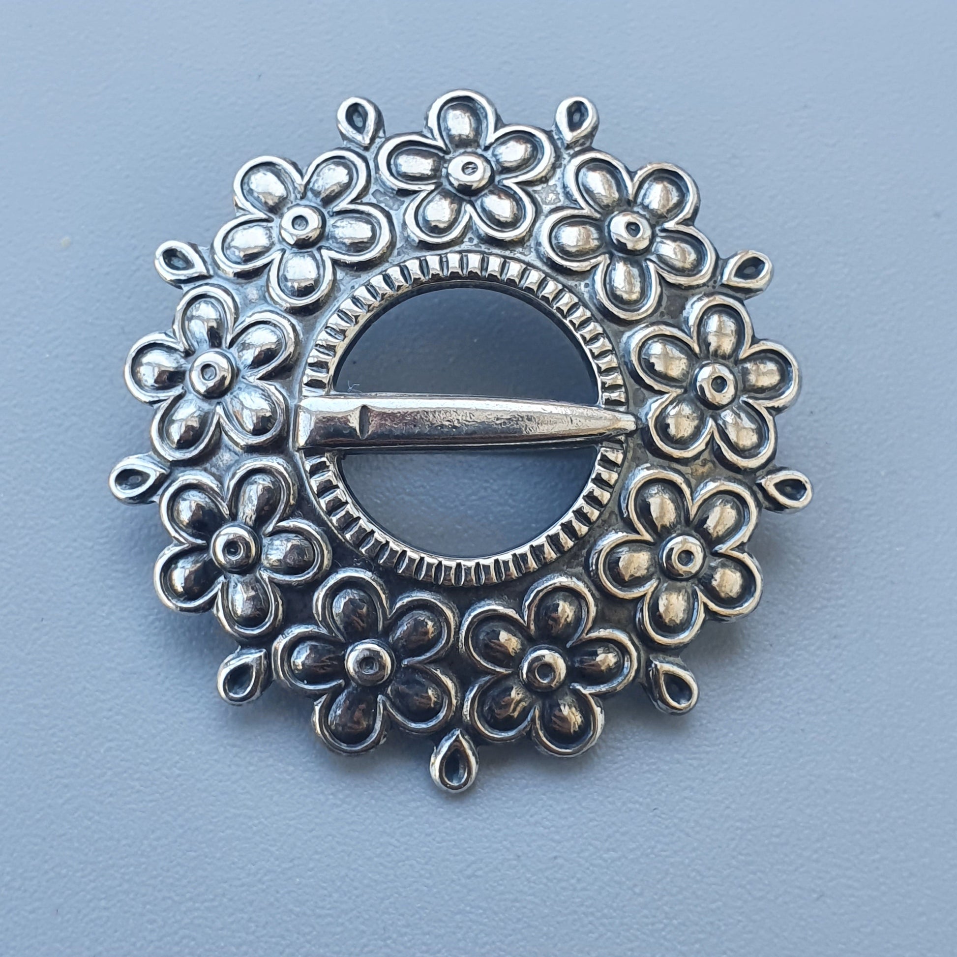 Circular silver brooch with floral design and central pin.