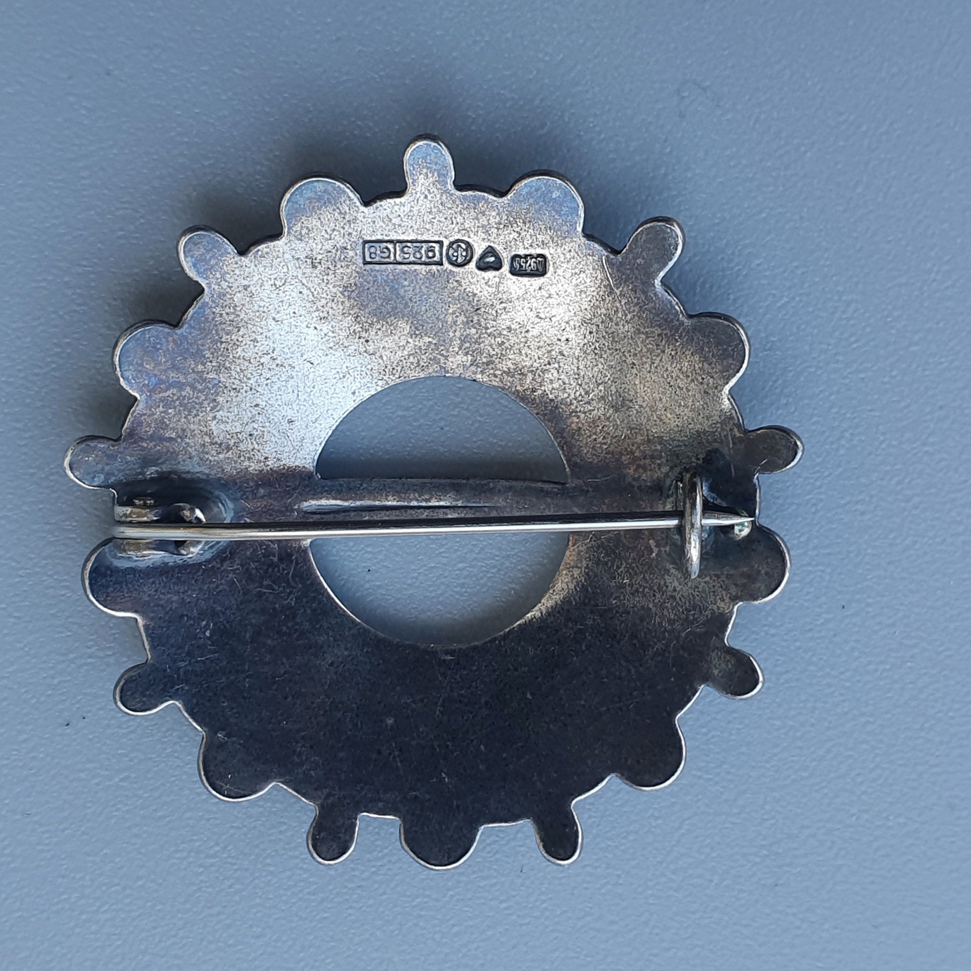 Silver gear-shaped brooch or pin with a central circular opening.