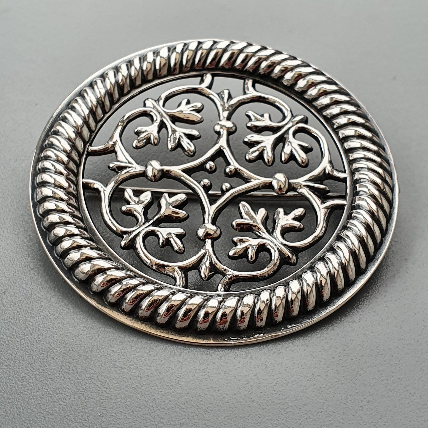 Ornate silver brooch with intricate floral and leaf designs encircled by a twisted rope border.