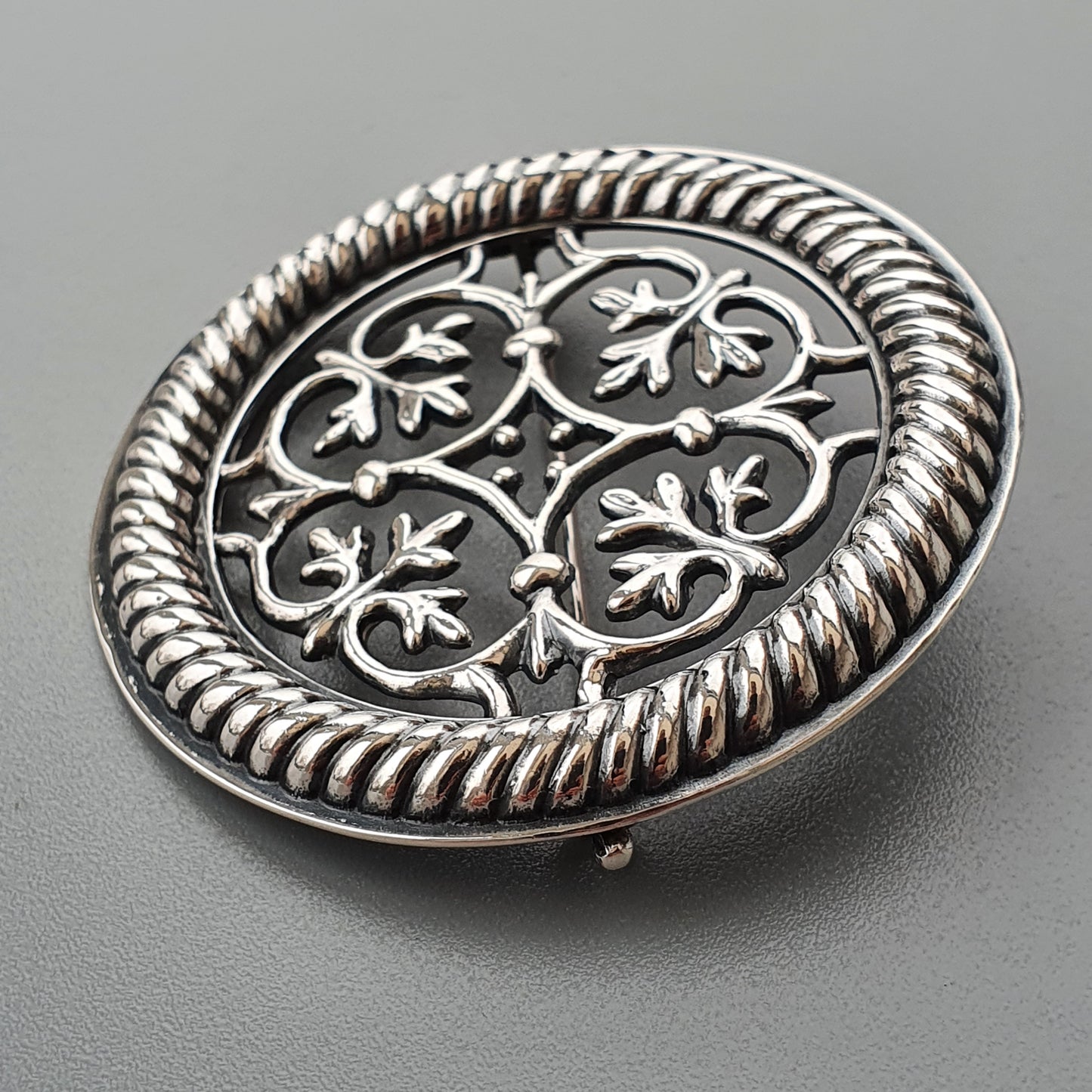 Ornate silver brooch with intricate floral filigree design and twisted rope border.