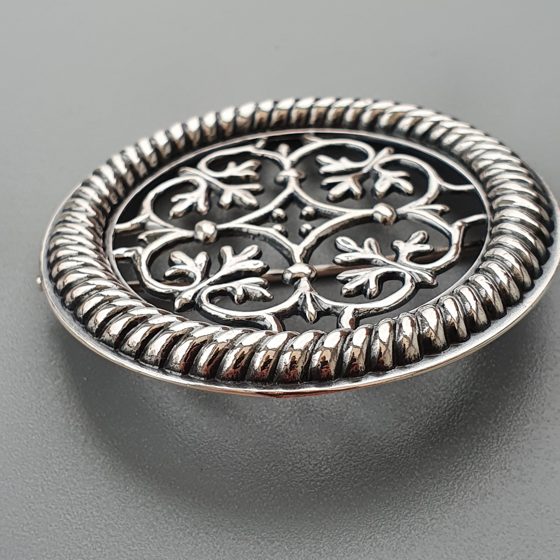 Ornate silver brooch or pendant with intricate floral filigree design and a twisted rope border.