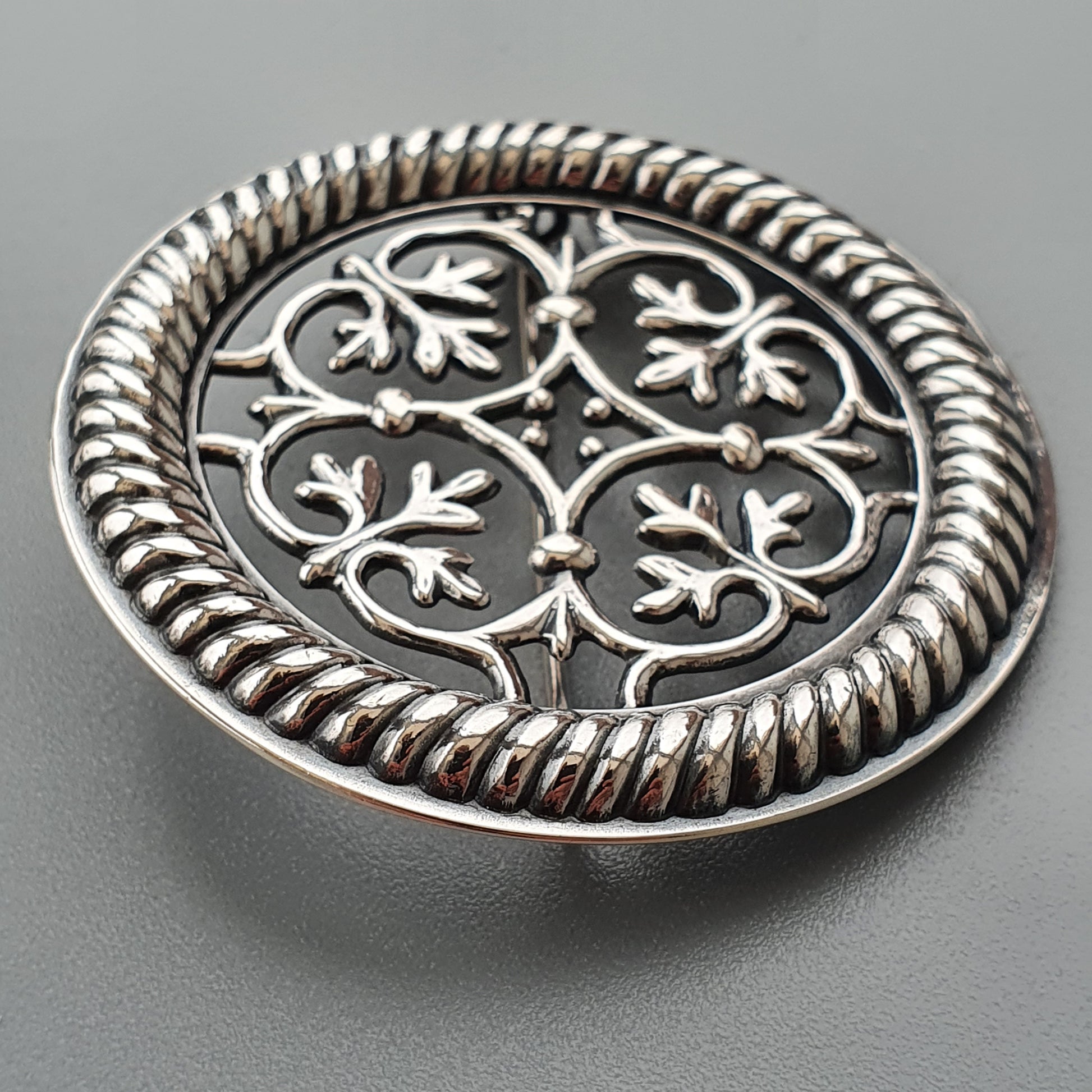 Ornate silver brooch with intricate floral filigree design and rope-like border.