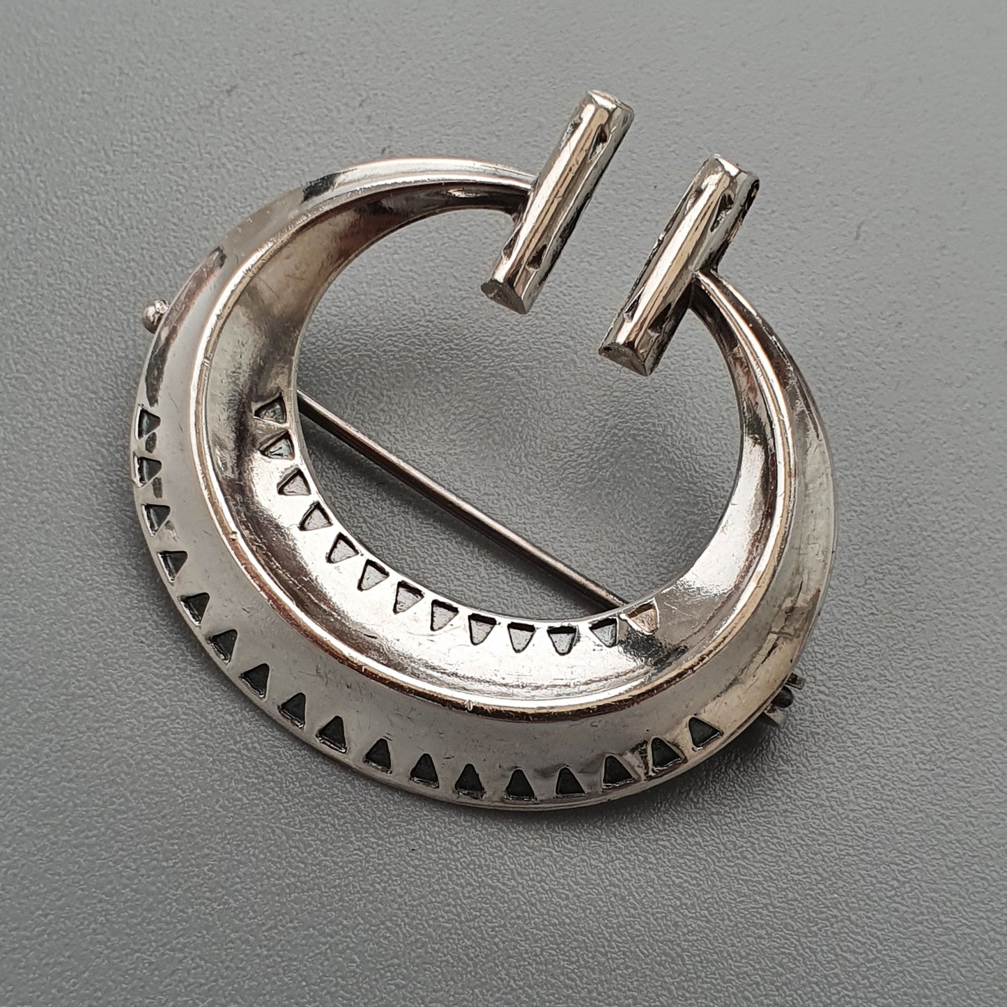 Crescent-shaped silver brooch or pendant with decorative triangular patterns and two protruding pins.