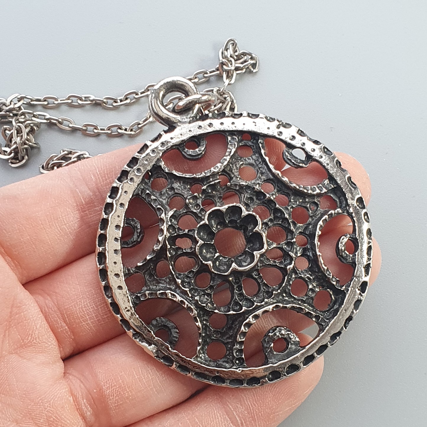 Ornate silver pendant with intricate floral filigree design on a reddish background.