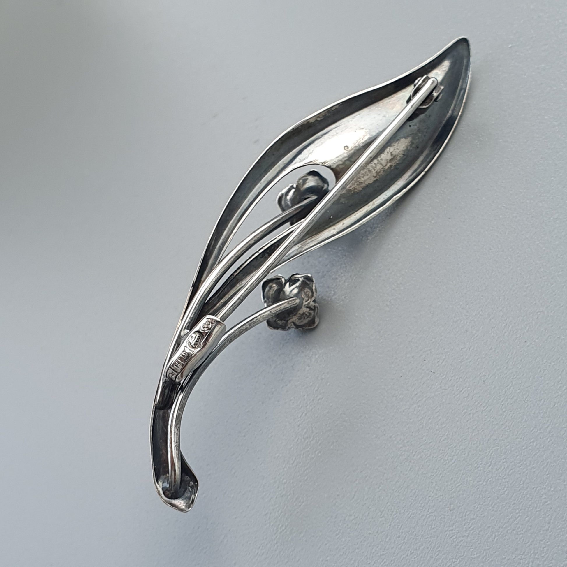 Sleek silver brooch shaped like an elongated leaf or feather with curved lines and small decorative elements.