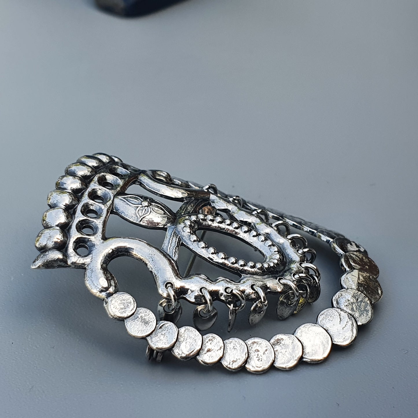 Ornate silver-colored metal jewelry piece with coin-like elements and intricate decorative patterns.