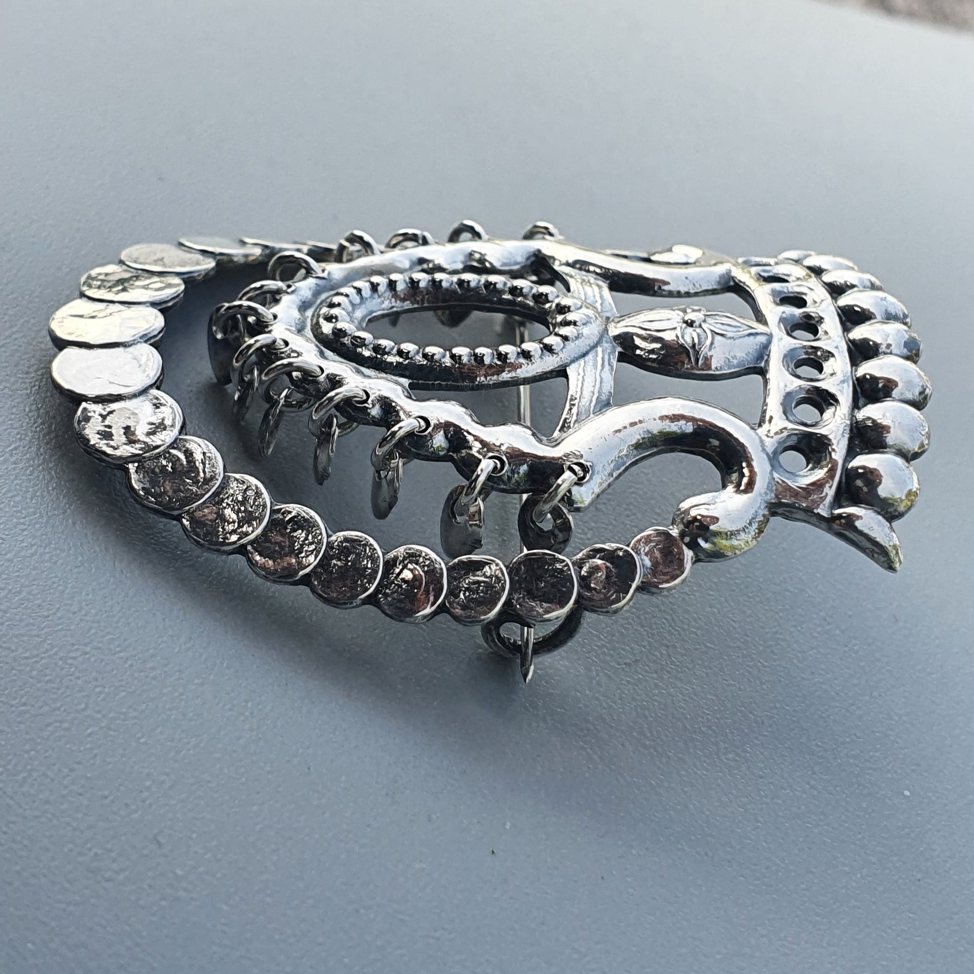 Metallic silver brooch or pin shaped like a stylized heart with intricate swirling and beaded designs.