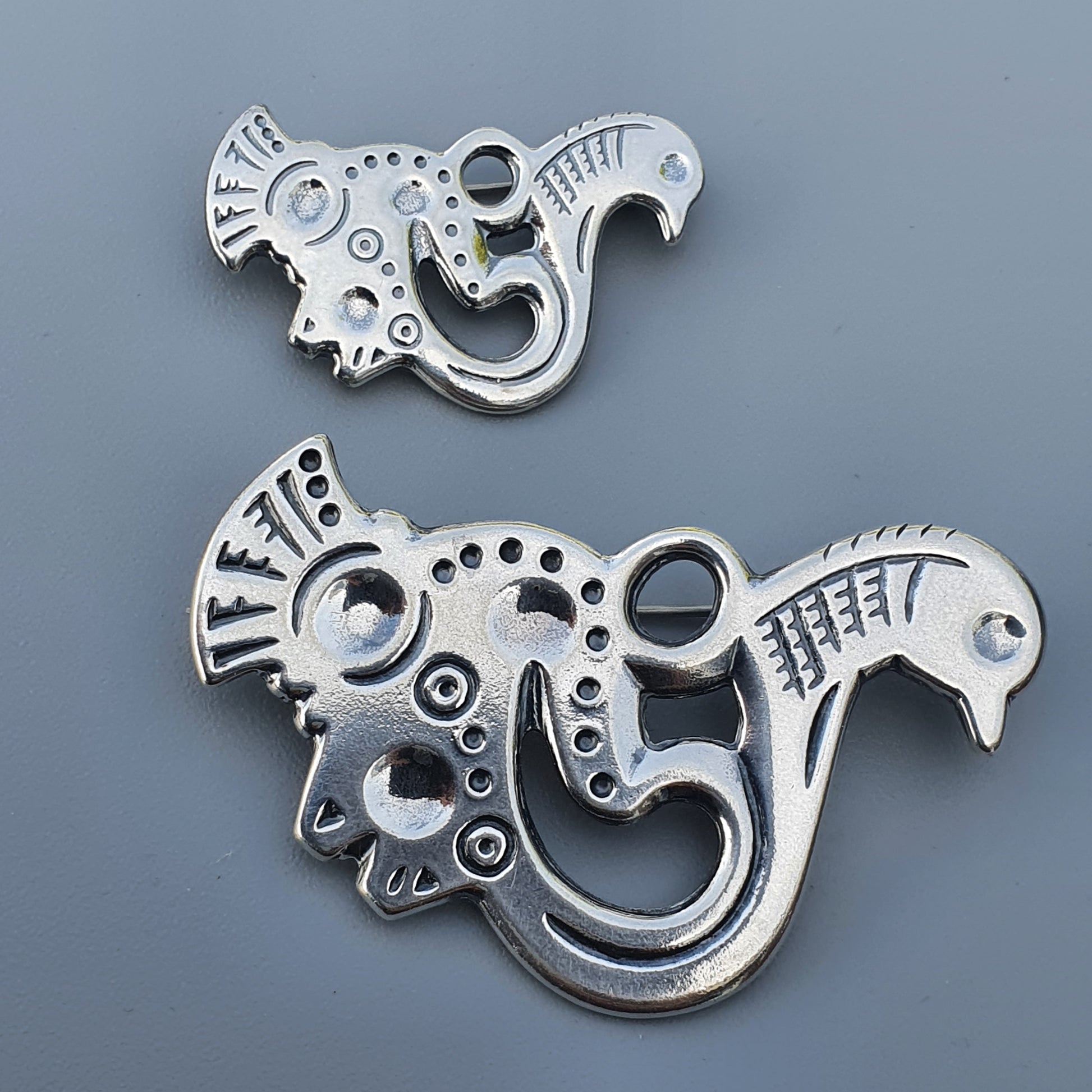 Stylized silver metal ornaments or pendants shaped like mythical bird-like creatures with intricate designs.