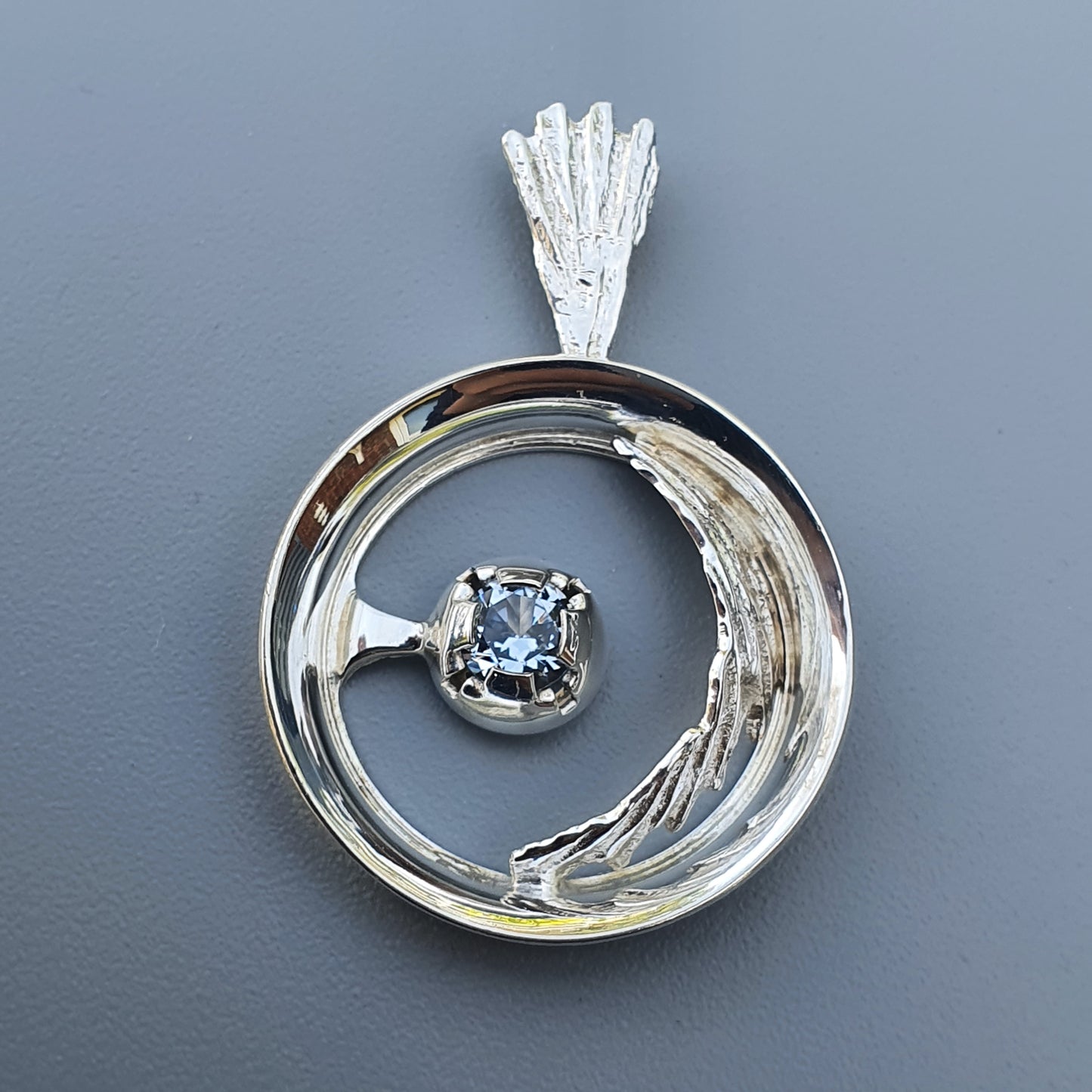 Circular silver pendant with a blue gemstone at its center.