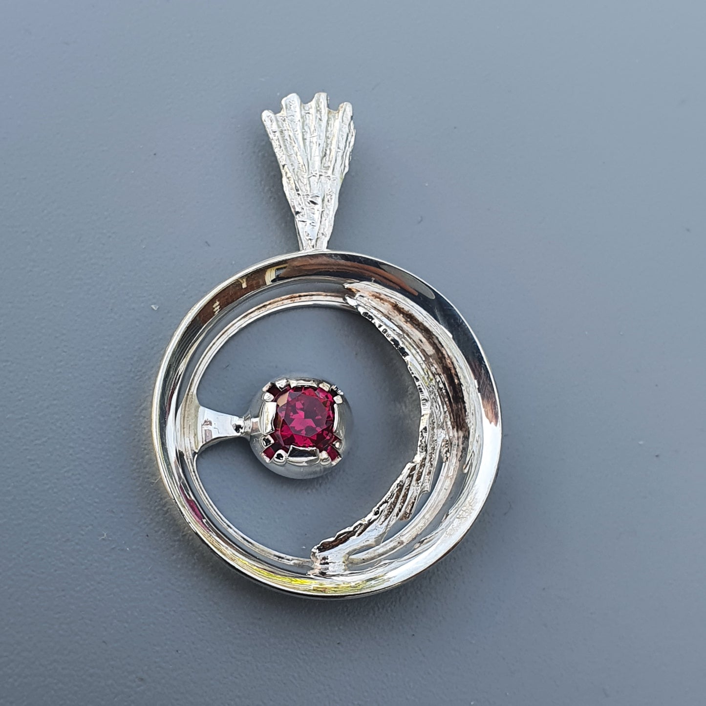 Circular silver pendant with a red gemstone at its center.