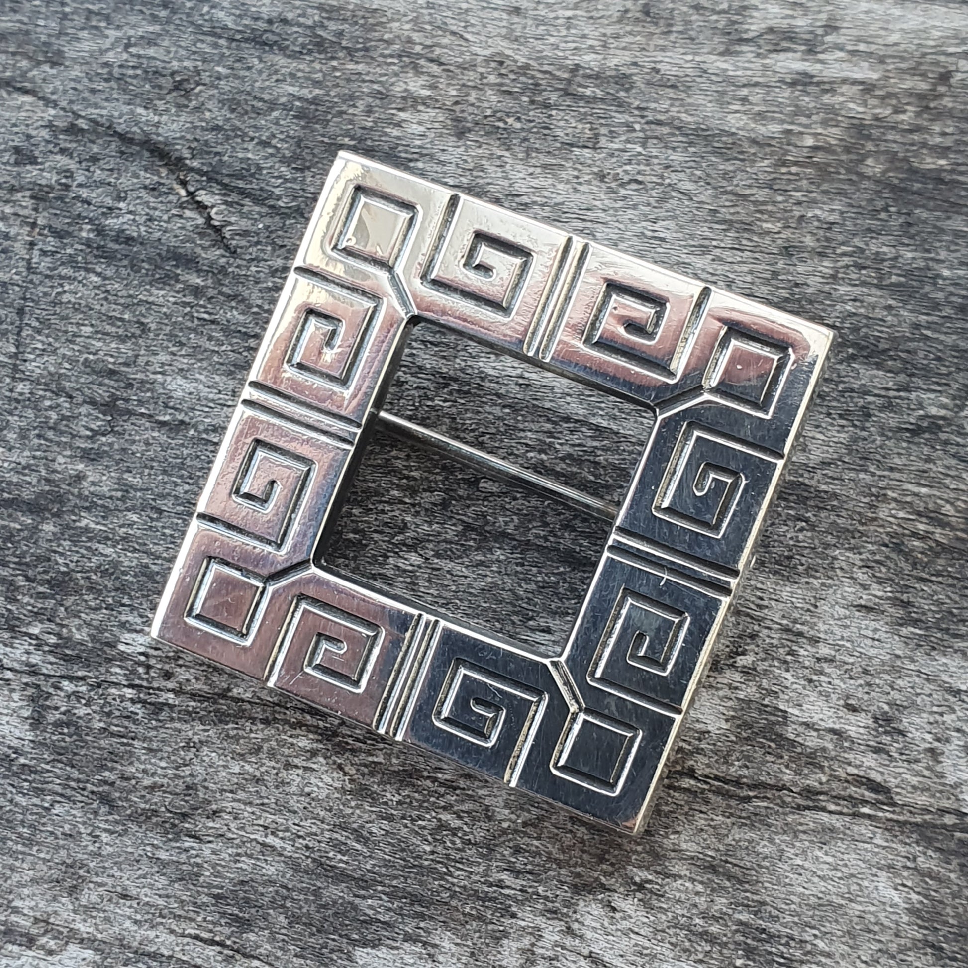 Square silver brooch with a Greek key pattern design.