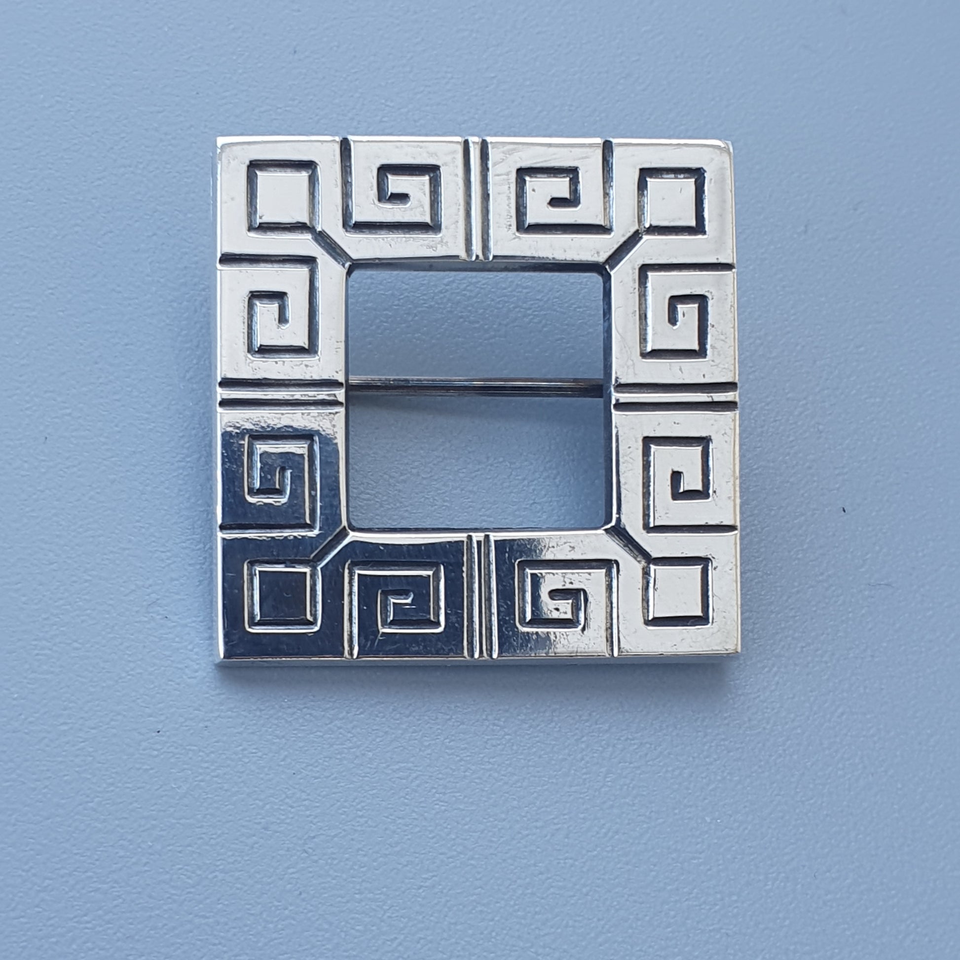 Square silver brooch or pin with a Greek key pattern around the edges.