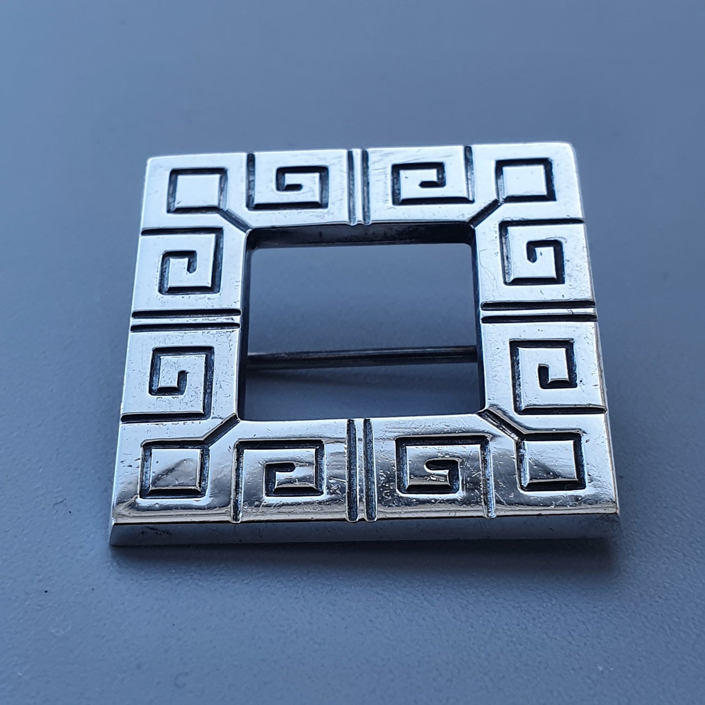 Square silver-colored belt buckle with a Greek key pattern design.