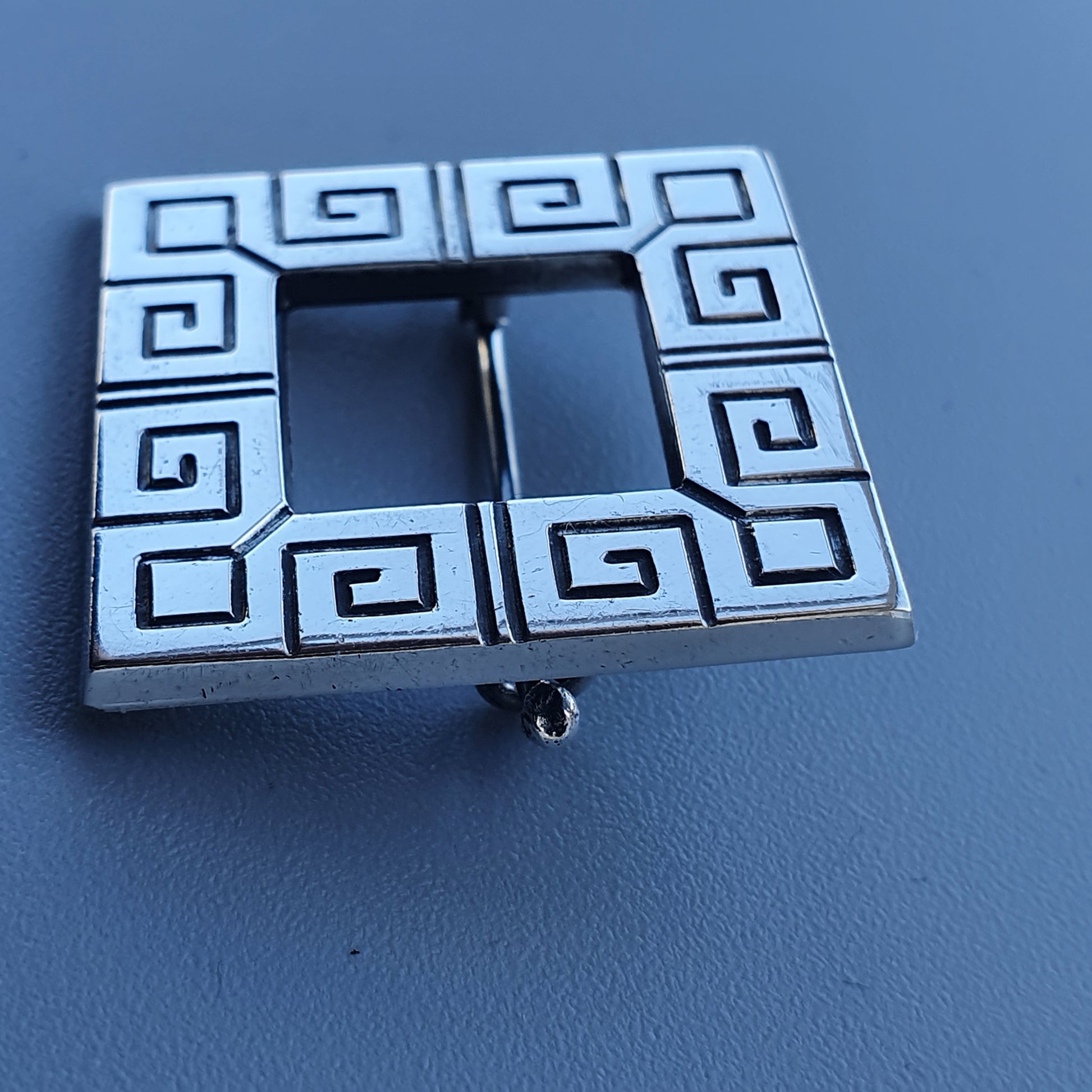 Square silver belt buckle with a Greek key pattern around the edges.