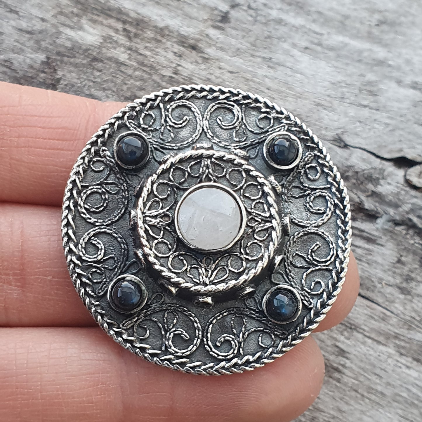 Ornate silver brooch with intricate swirling patterns, a central white stone, and four blue stones.