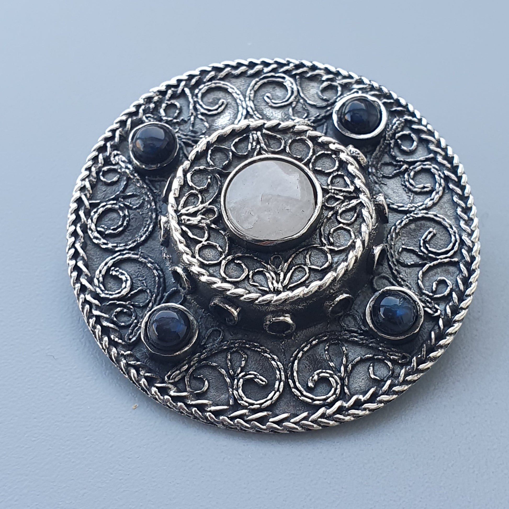 Ornate silver brooch with a central white stone surrounded by blue gemstones and intricate filigree patterns.