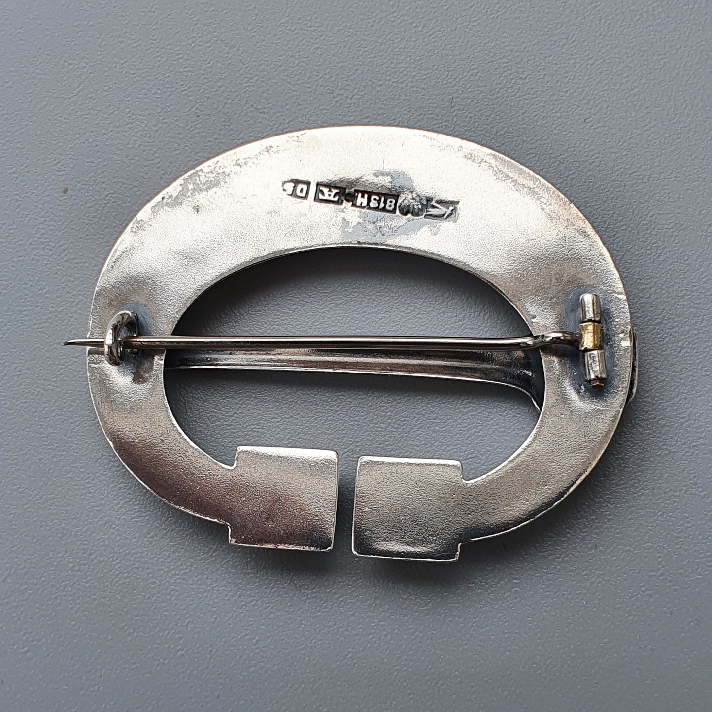 Silver oval brooch with a horizontal pin and geometric cutouts at the bottom.