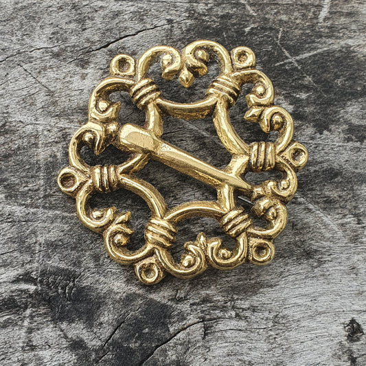 Ornate golden brooch with intricate swirling patterns and a central cross-like design.