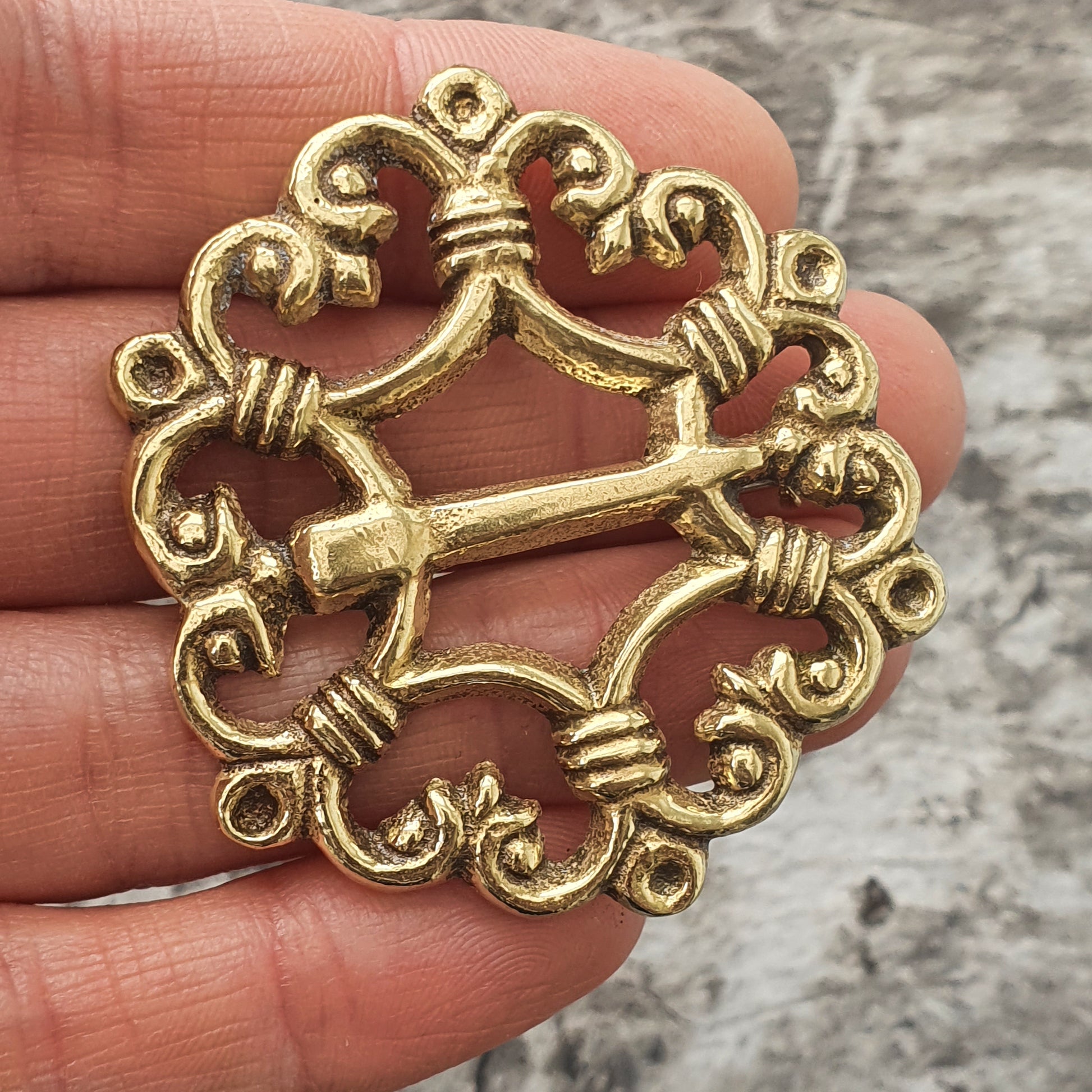 Ornate gold-colored metal brooch or pin with a circular design featuring scrollwork and a central bar.