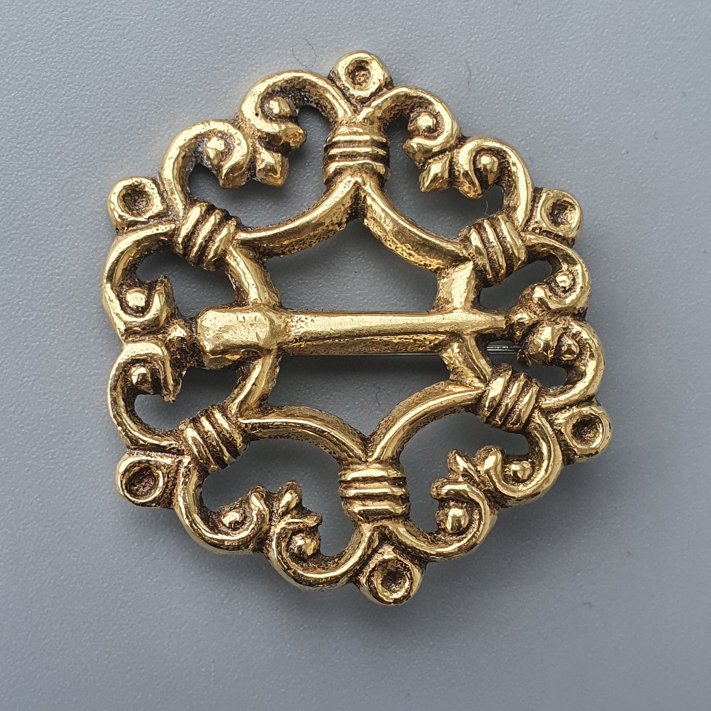 Ornate gold-colored circular brooch or clasp with intricate scrollwork and a central bar.