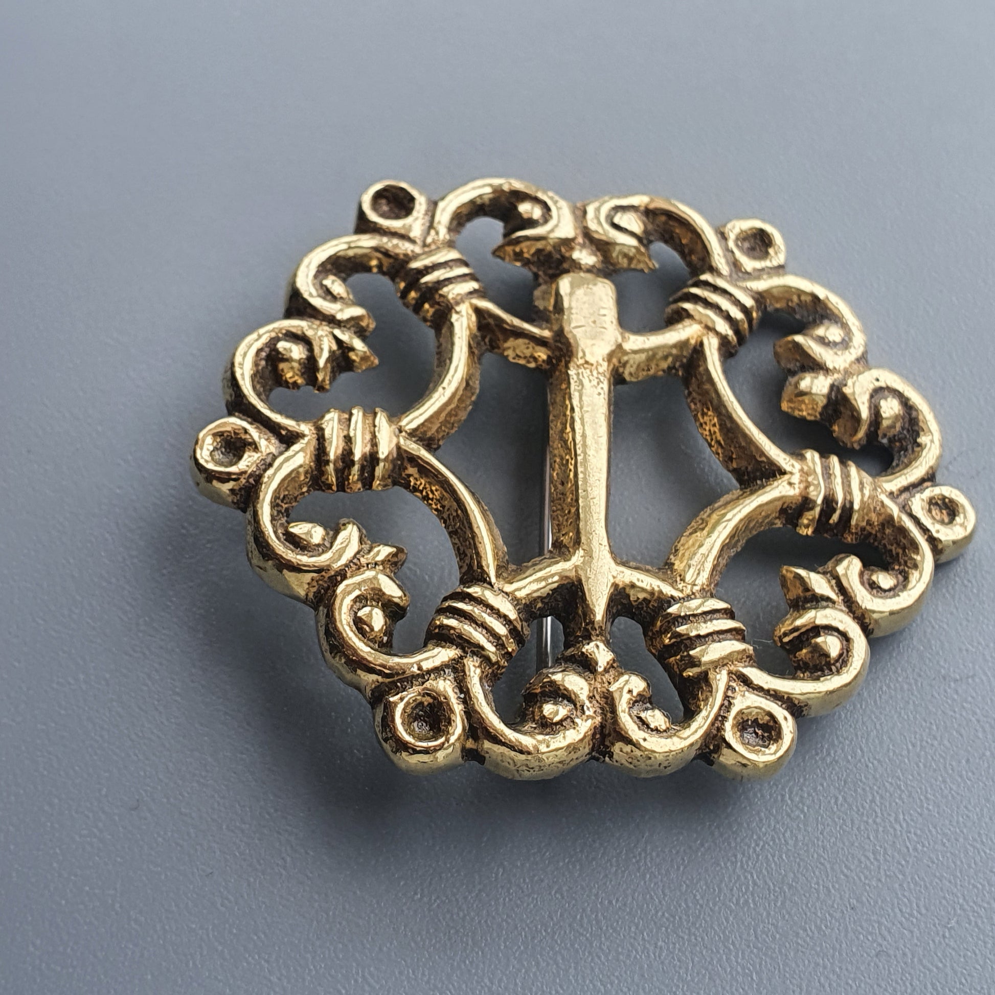 Ornate gold-colored metal brooch or pendant with intricate scrollwork and a central cross design.