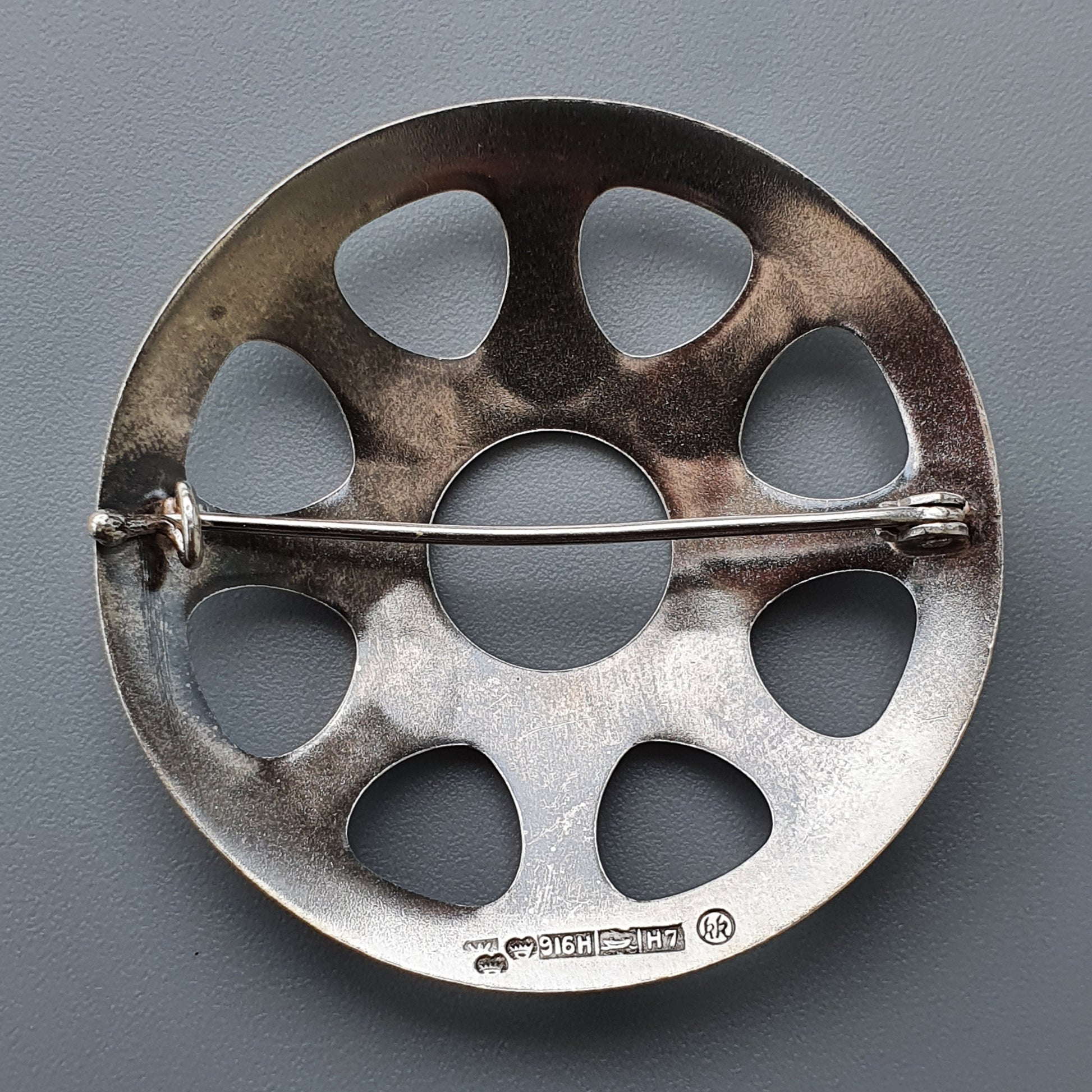 Circular metal brooch or pin with multiple teardrop-shaped cutouts and a horizontal bar across the center.