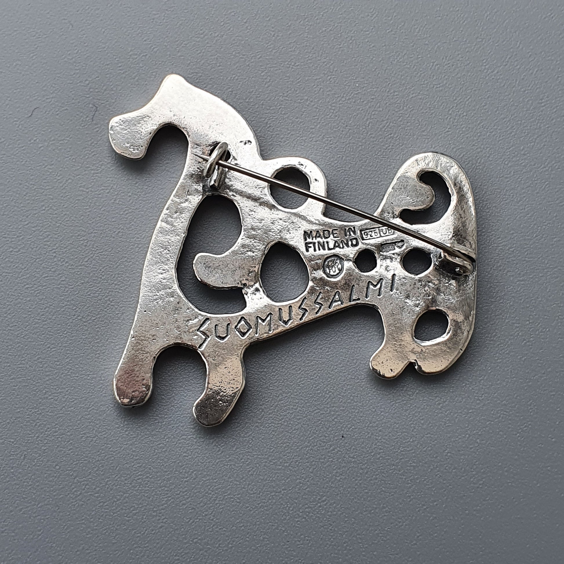 Silver brooch shaped like a stylized dog with cutout designs and ’SUOMI FINLAND’ engraved on it.