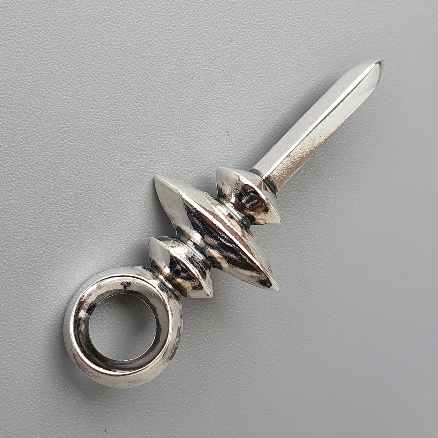 Metallic pendant or charm with a pointed blade-like shape and circular loop at one end.