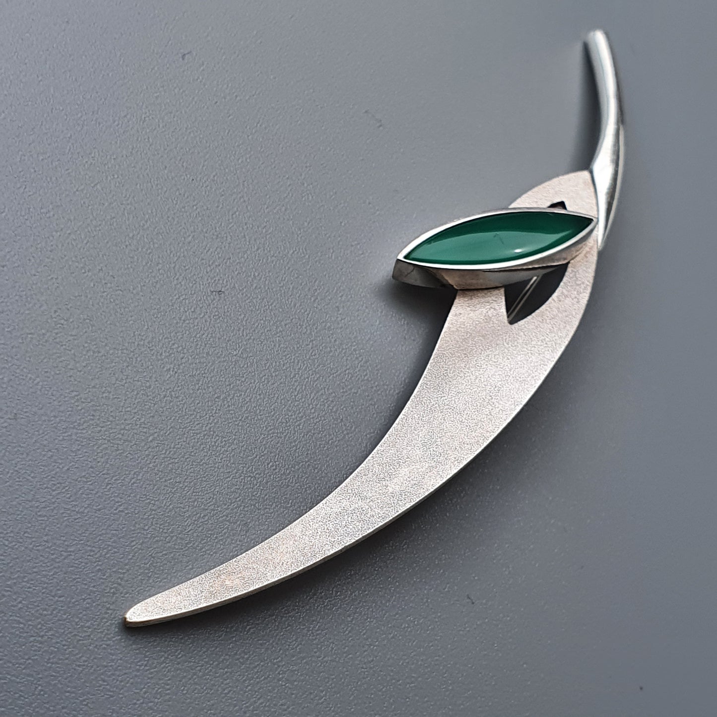 Sleek silver brooch with a curved shape and green gemstone accent.