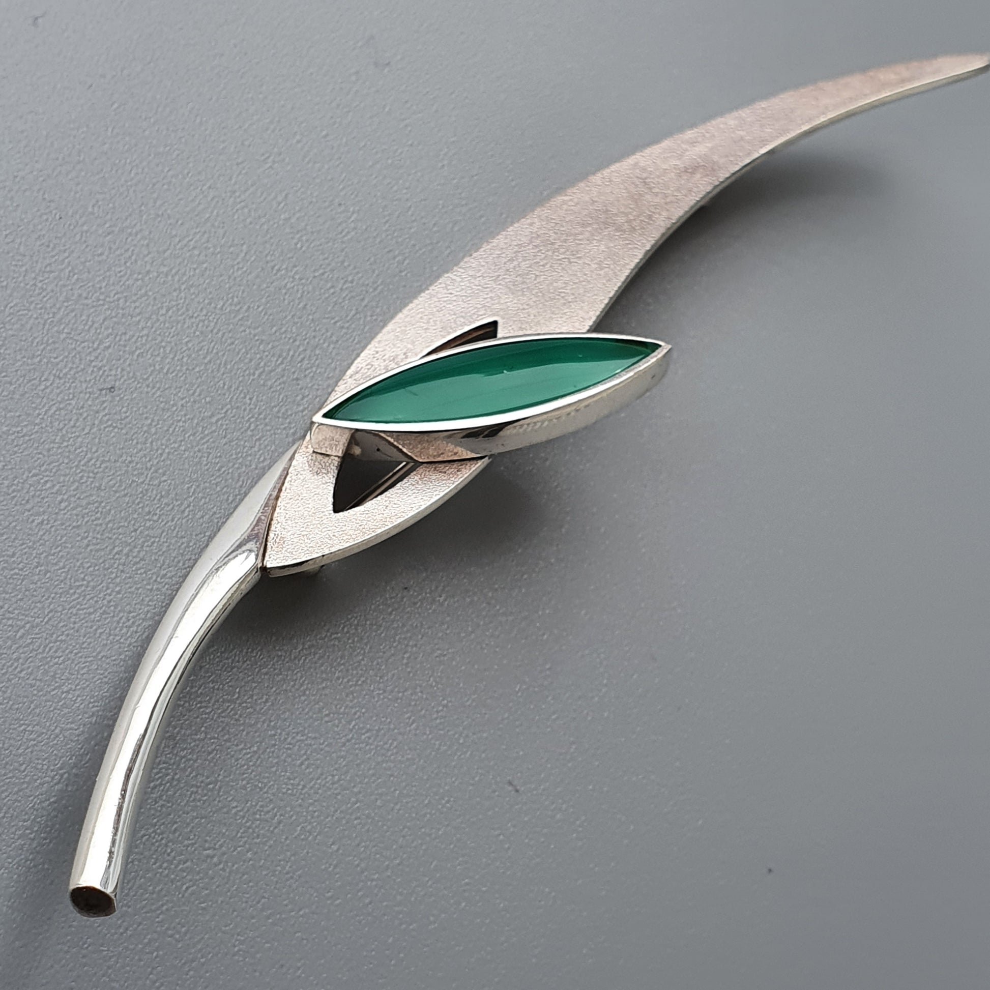 Sleek silver brooch with a curved stem and leaf-like shape containing a green enamel accent.