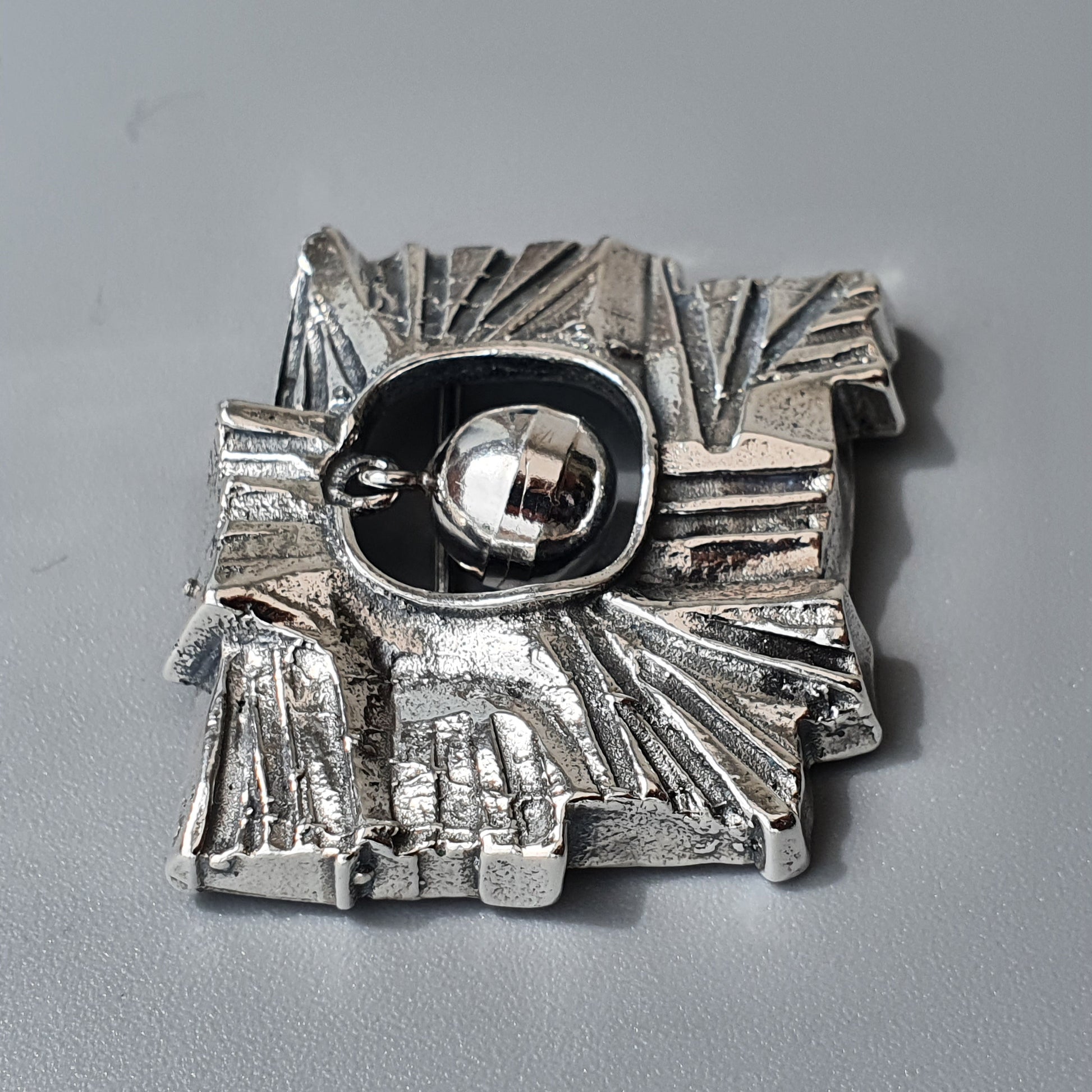 Silver brooch or pendant with an abstract sunburst design and a central black oval containing a shiny sphere.