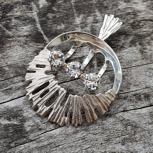 Circular silver brooch with three diamonds and a fan-like design.