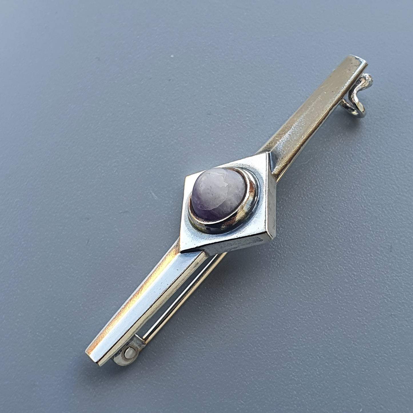 Metallic tie clip with a diamond-shaped centerpiece containing a round purple stone.