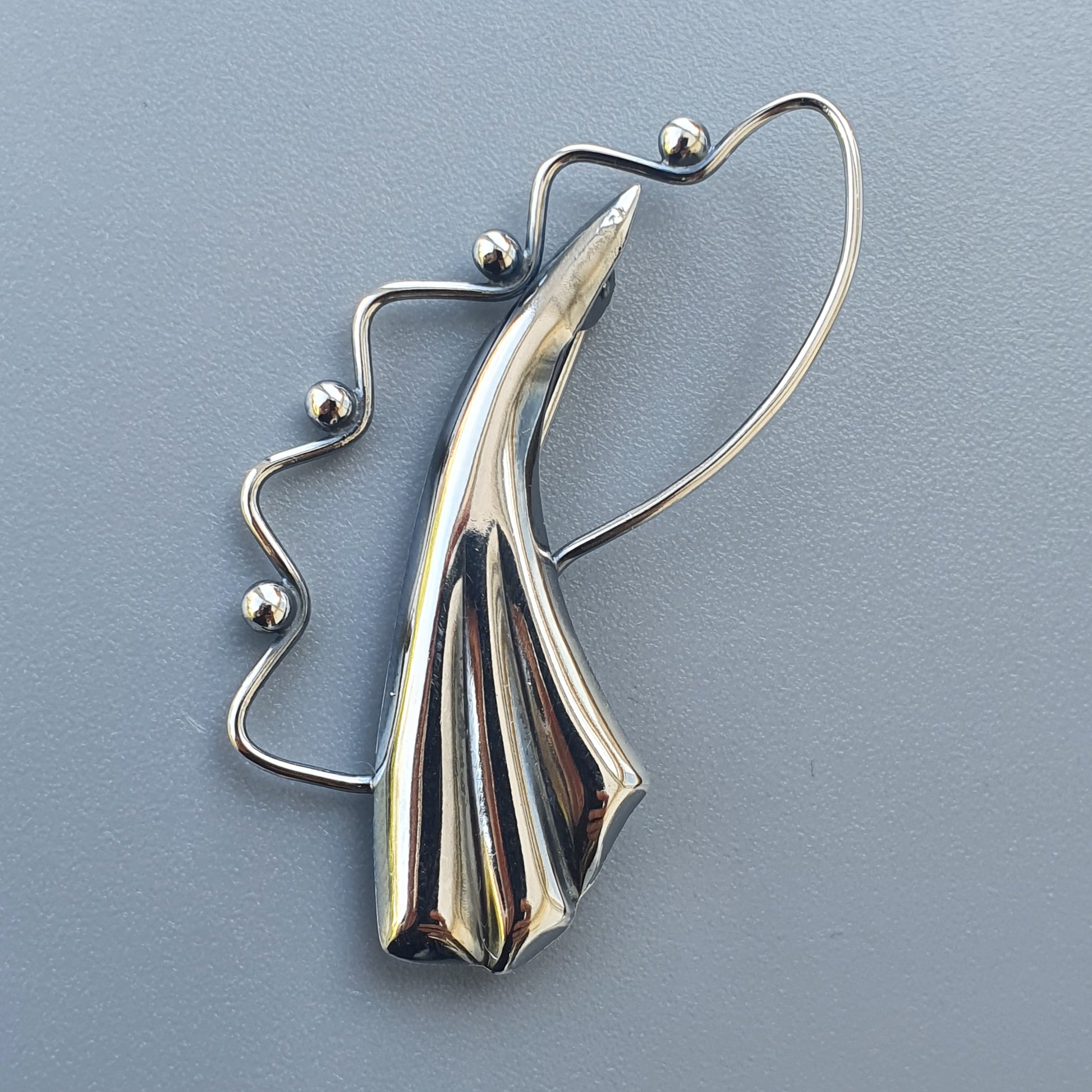 Sleek silver brooch or pendant with a curved, abstract wing-like design and beaded wire accent.