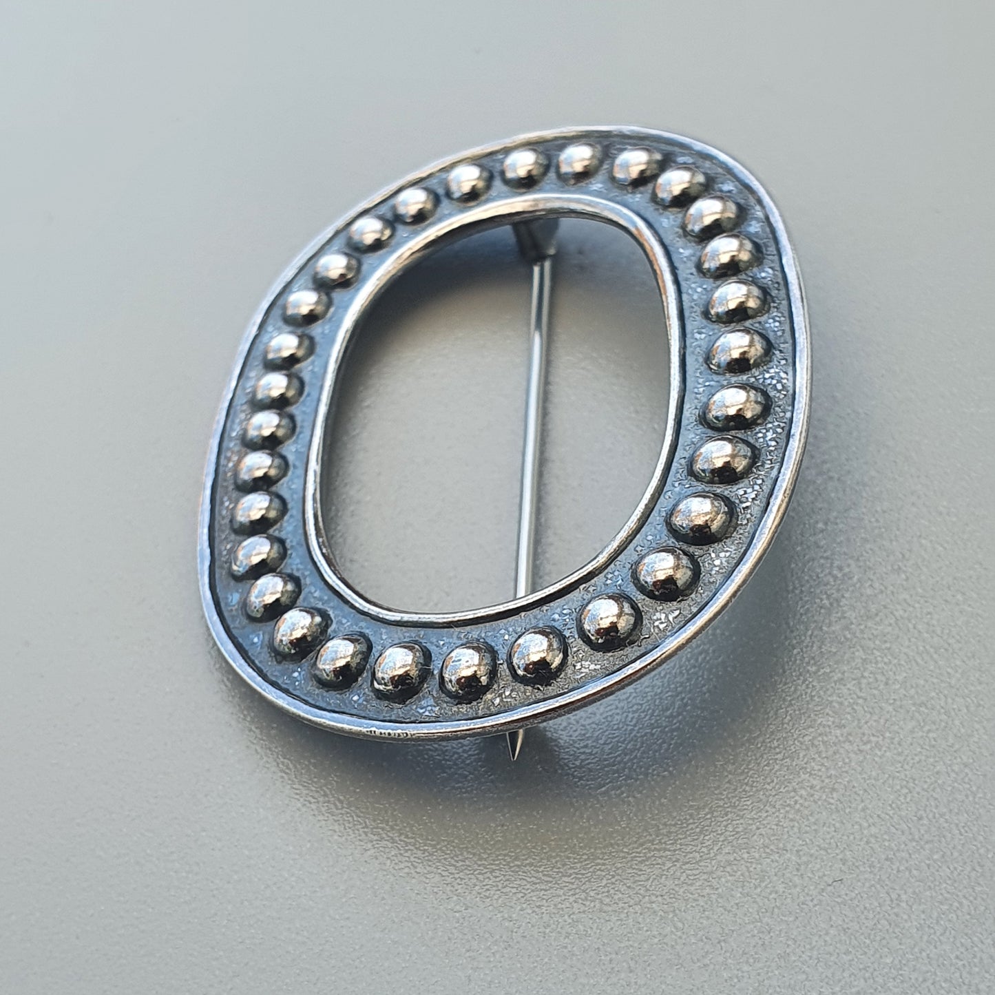 Oval-shaped silver brooch or pin with beaded edge detailing.