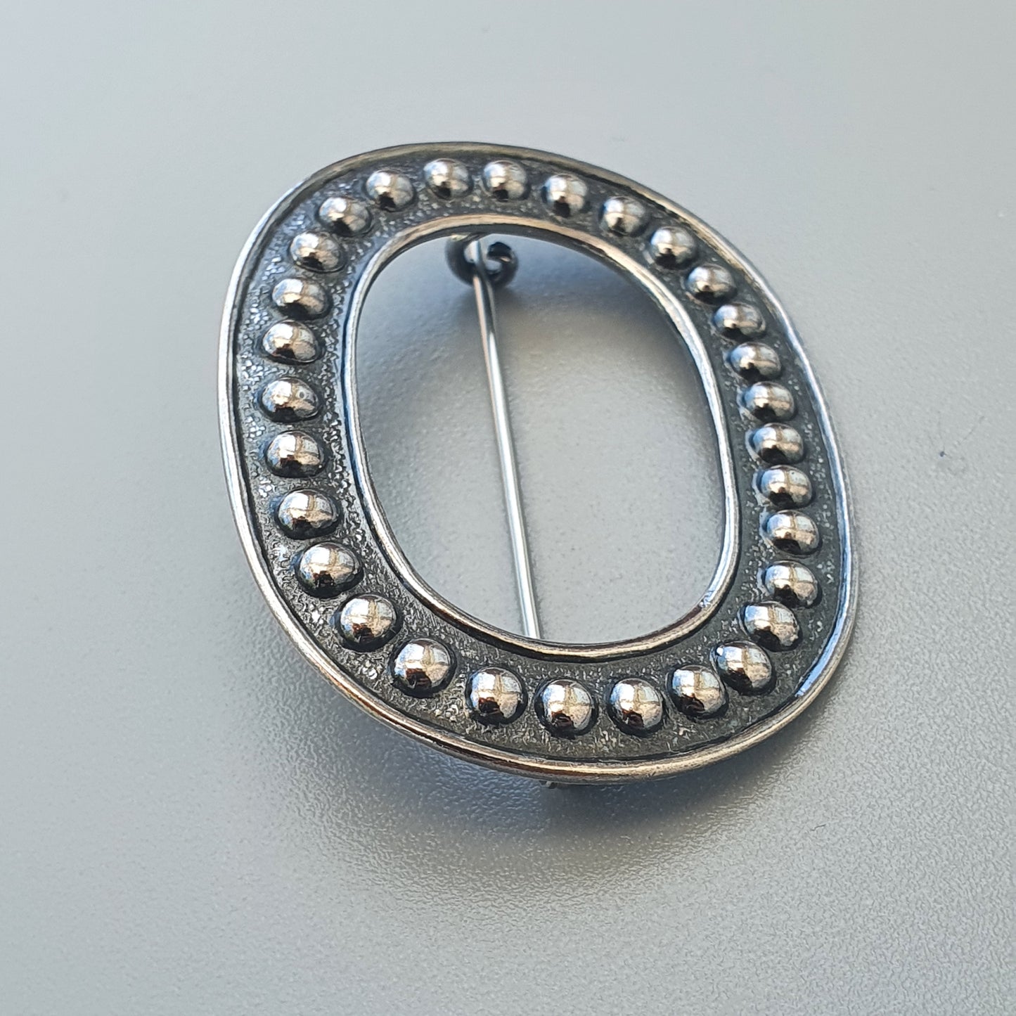 Oval-shaped silver brooch or pin with beaded edge decoration.