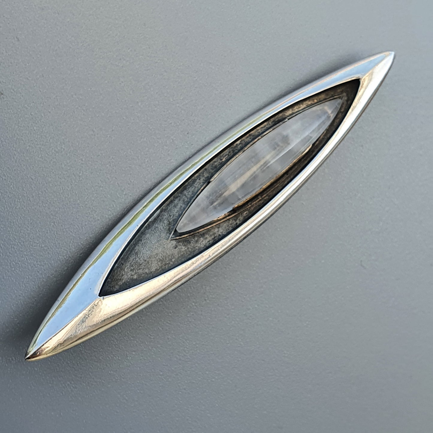 Sleek, elongated silver brooch or pendant with an eye-shaped design and a central clear gemstone or crystal.