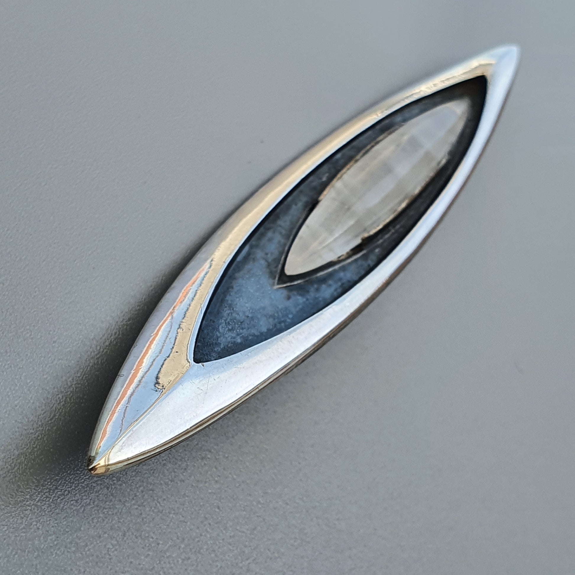 Sleek, eye-shaped metallic brooch or pendant with layered silver, blue, and gold tones.