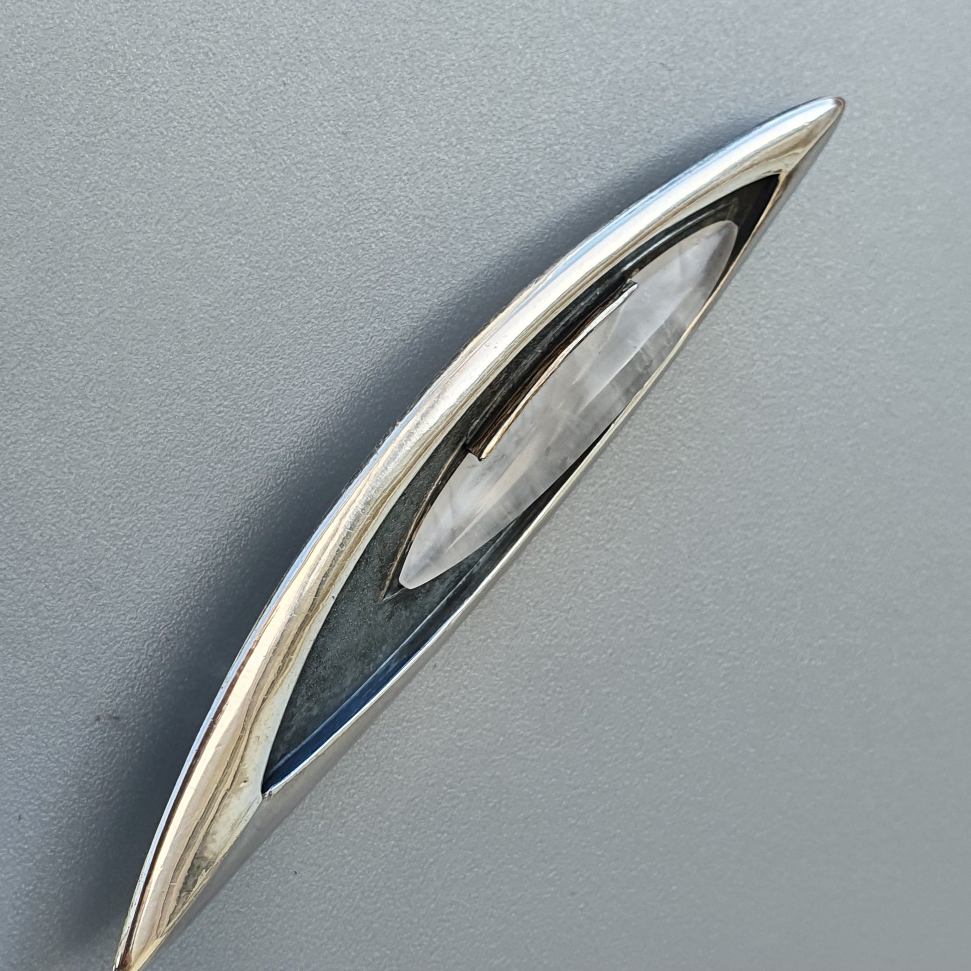 Sleek, elongated silver brooch or pin with a curved shape and inset oval gemstone or crystal.