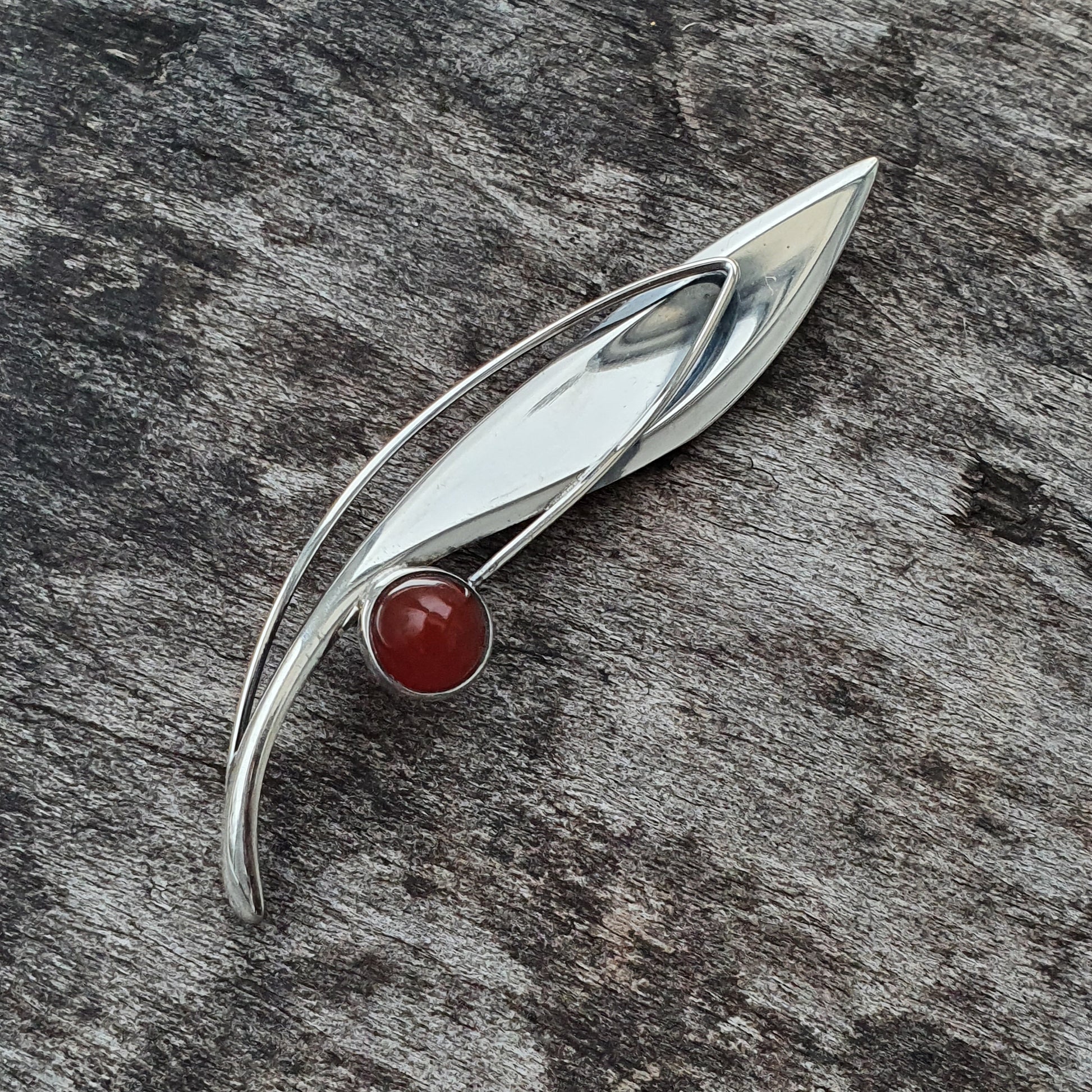 Sleek silver brooch with a curved leaf-like design and a red gemstone accent.