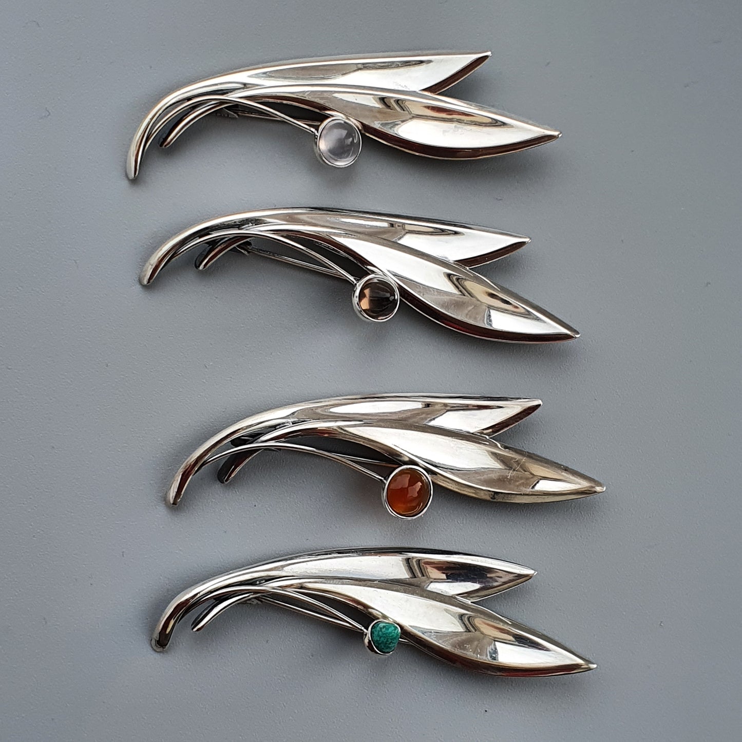 Sleek, silver brooches shaped like stylized leaves or feathers, each adorned with a different colored gemstone.