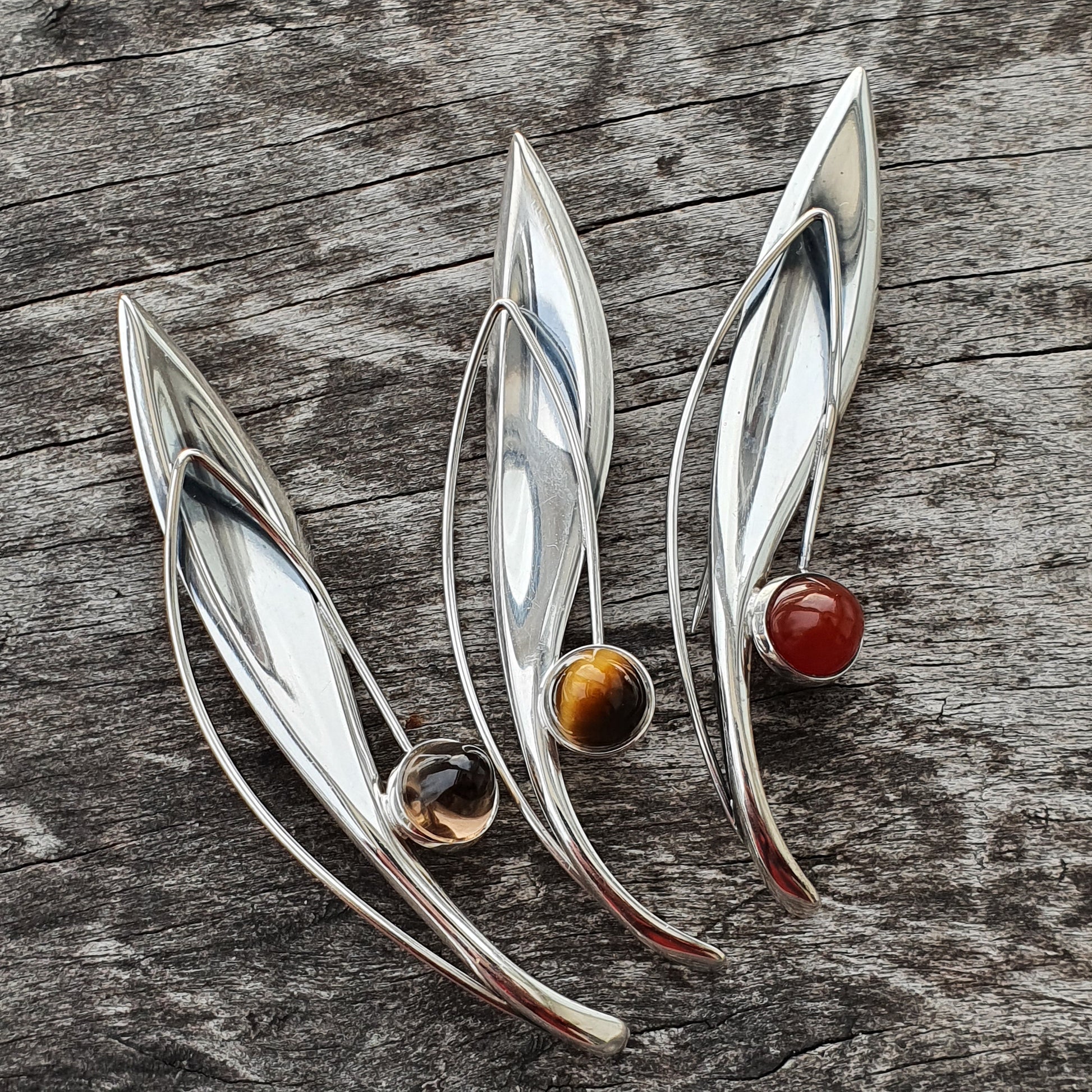 Three sleek, leaf-shaped silver brooches, each adorned with a colorful gemstone bead.