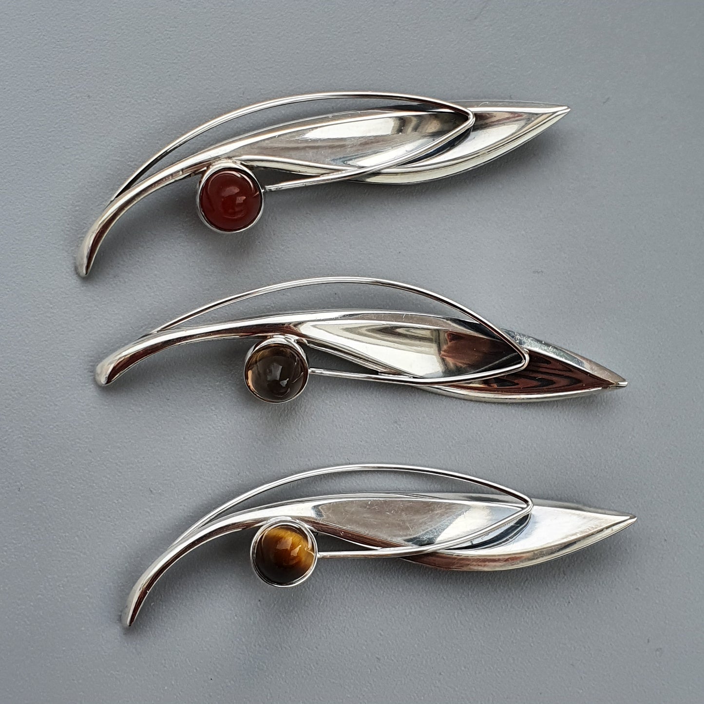 Three sleek, leaf-shaped silver brooches, each adorned with a colorful gemstone.