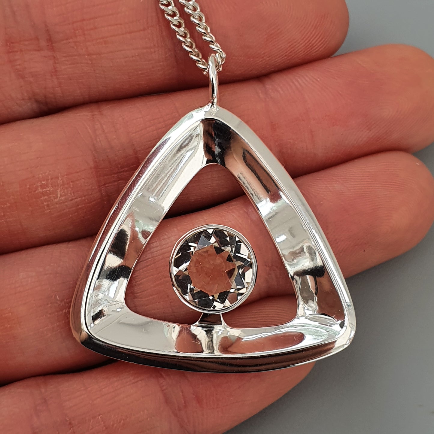 Silver triangular pendant with a circular gemstone in the center, suspended on a chain.