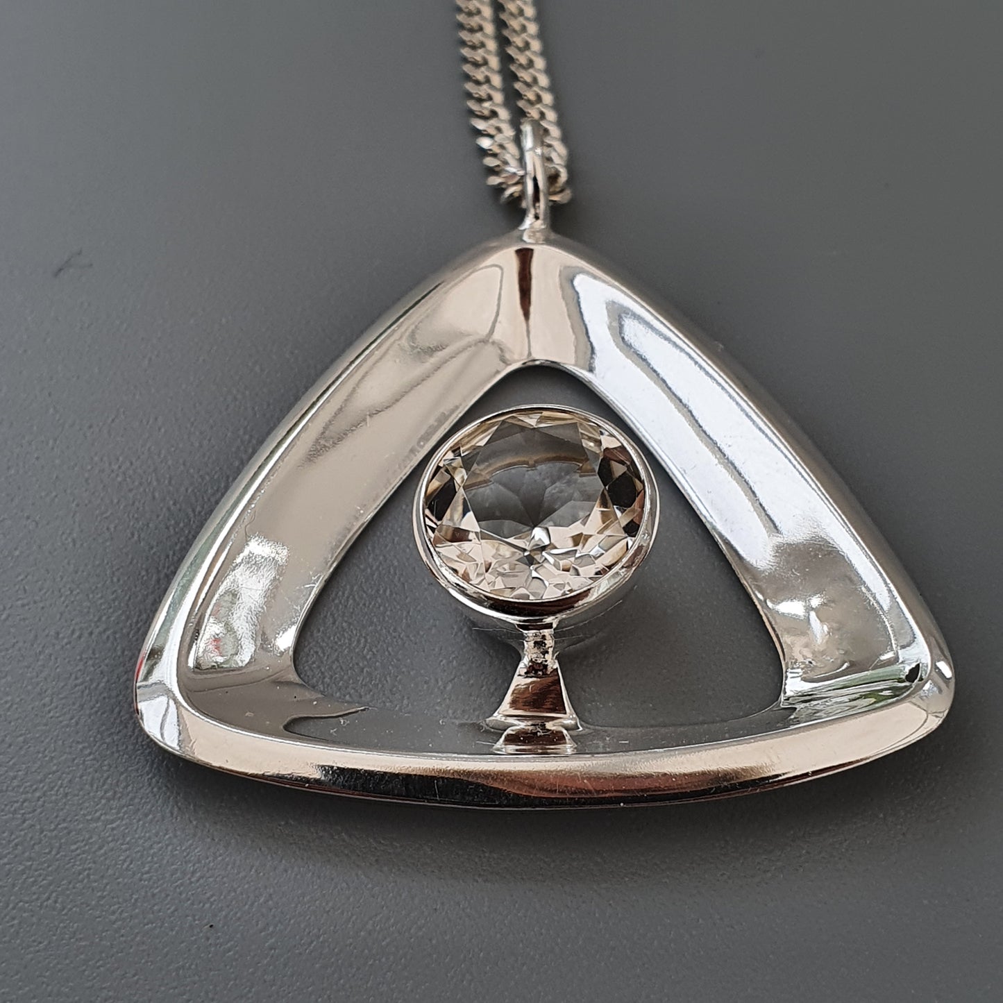 Triangular silver pendant with a round crystal or gemstone at its center, suspended on a chain.