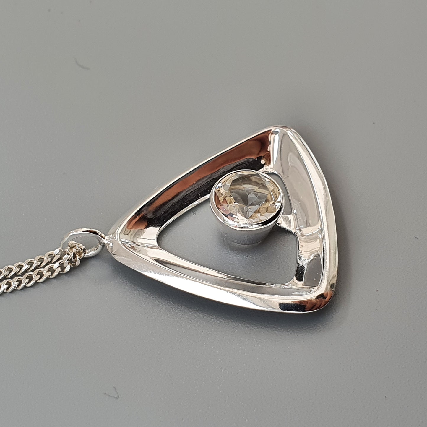 Triangular silver pendant with a clear gemstone in the center, attached to a chain.