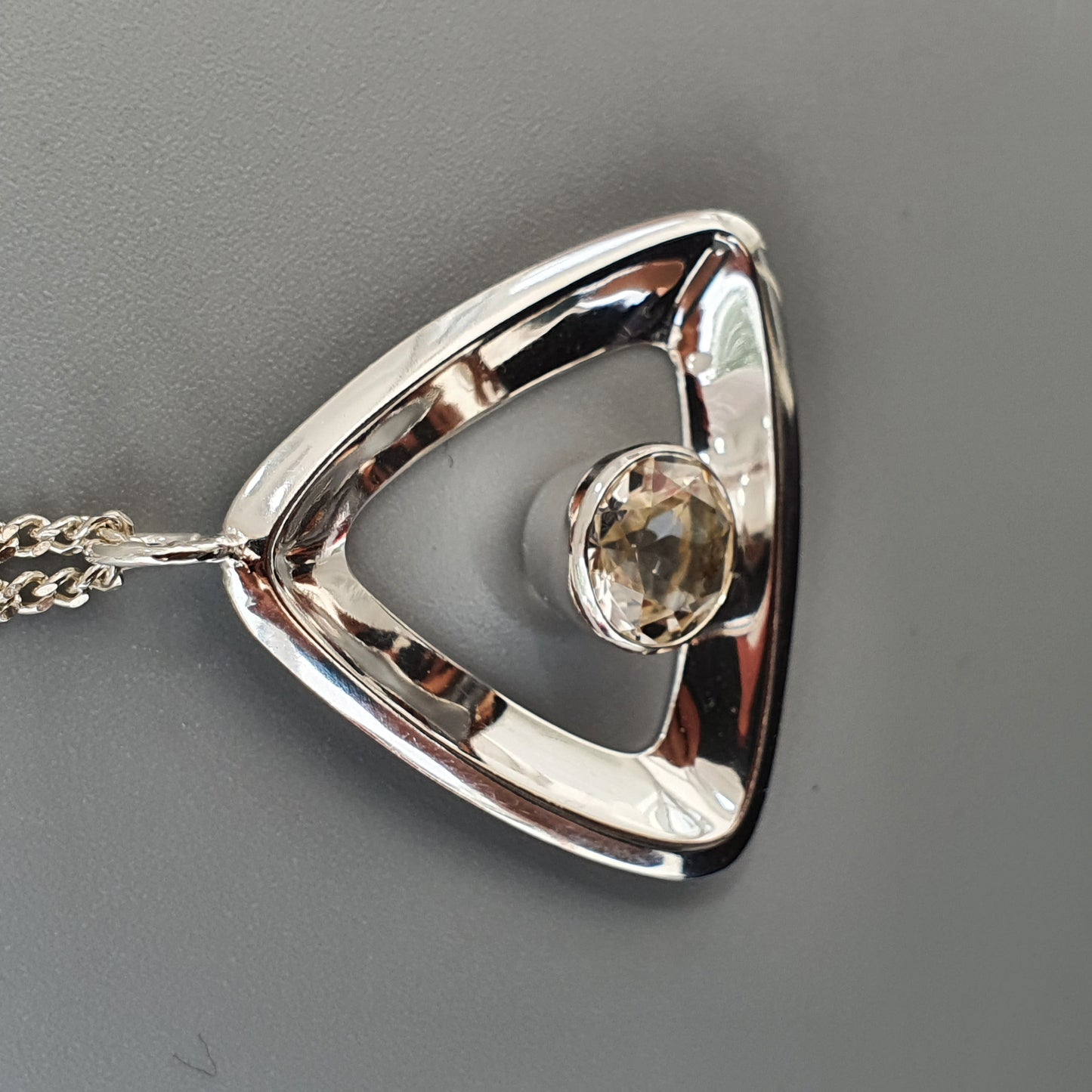 Silver triangular pendant with a clear gemstone in the center, attached to a chain.