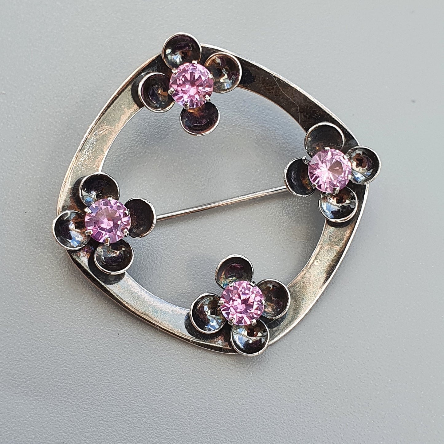 Circular silver brooch with four flower-shaped designs featuring pink gemstones.